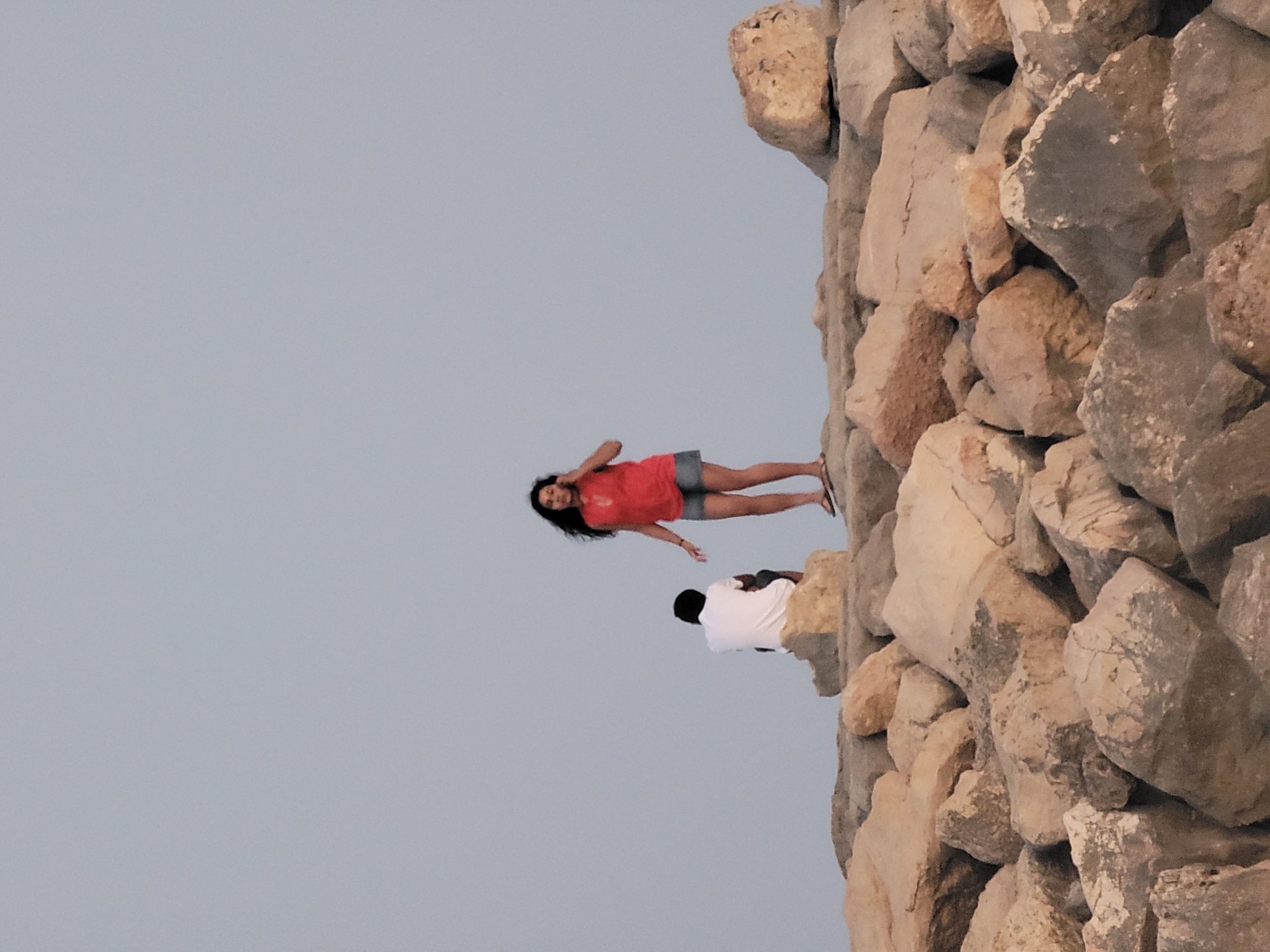 Two individuals are positioned on a rocky embankment against a pale, overcast sky. The person on the left is a dark-skinned male wearing a white t-shirt and dark shorts, seated with his back to the camera. The person on the right is a fair-skinned female with long, dark hair, standing and looking to her left. She is wearing a red top and denim shorts, and appears to be holding her left hand to her ear, possibly talking on a phone or listening. Both are on separate large rocks on the embankment. The embankment is composed of numerous large, light-colored rocks. This scene is consistent with the rocky shoreline found along the Quarteira coastline in Portugal, specifically areas like the breakwaters or sections of the promenade featuring natural or reinforced rock formations.