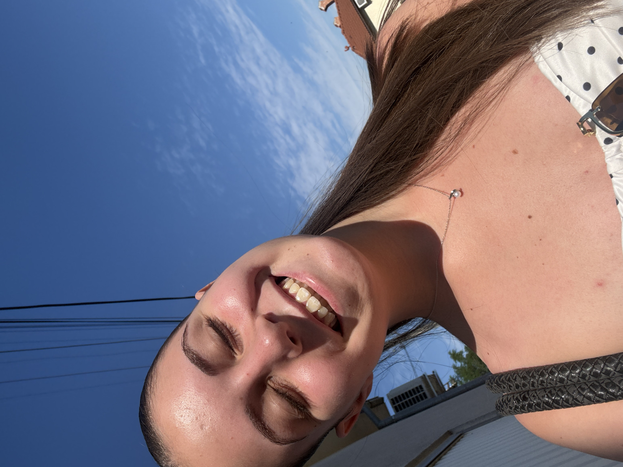 The media file is a selfie of a young woman outdoors. She has long, dark brown hair and is smiling with her eyes closed. She is wearing a white top with black polka dots and a black braided bracelet. A thin silver necklace with a small pearl pendant is visible around her neck. In the background, the sky is bright blue with some white clouds, suggesting it is a sunny day. Some power lines are visible across the sky, and a building with an air conditioning unit is in the distance. The image is taken from a low angle, looking up at the woman against the sky. The overall impression is one of joy and freedom.