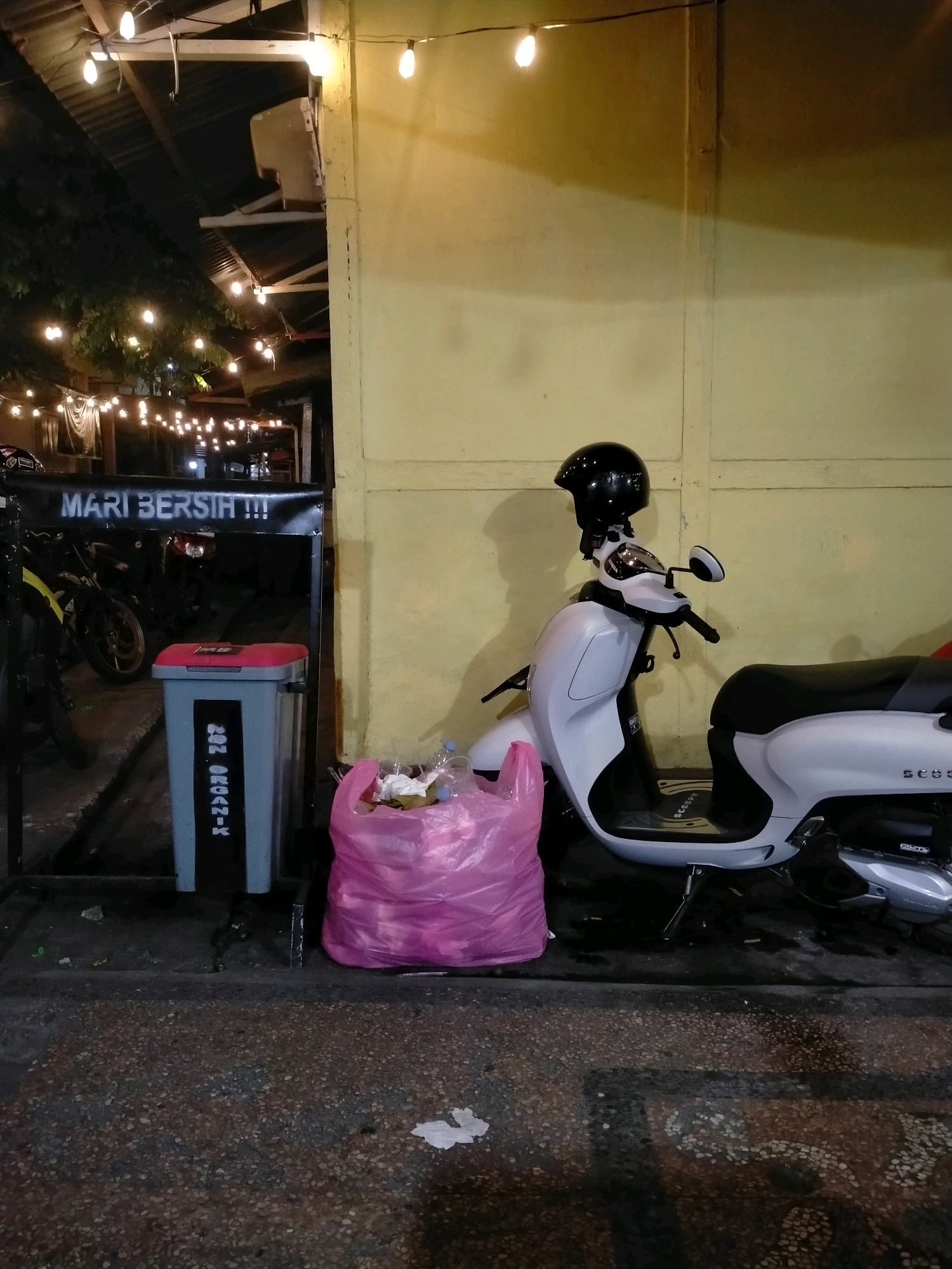An outdoor scene at night in Ambon, Indonesia, shows a light-colored scooter parked in front of a yellow wall. A black helmet rests on the scooter's handlebars. To the left of the scooter, a gray trash can with a red lid stands next to a black metal frame. The trash can has text on it, including "NON ORGANIK." A large, pink plastic bag filled with trash is placed on the ground to the right of the trash can. Overhead, string lights are visible, some illuminated, suggesting a commercial or outdoor dining area. The ground is paved with a textured material.