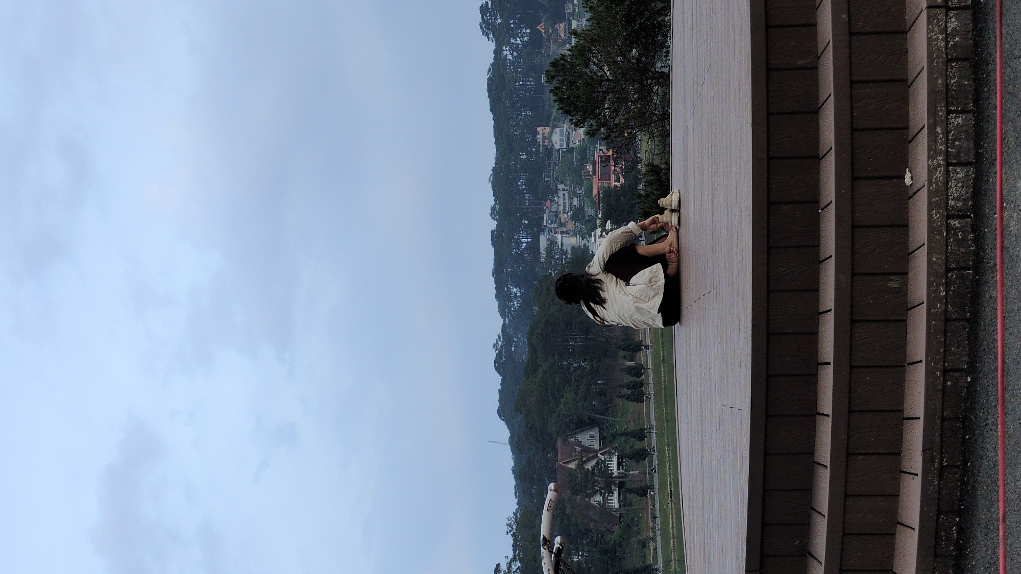 A woman sits on a wooden ledge overlooking a cityscape in Xuân Hương - Đà Lạt, Vietnam. She is curled up, facing the city, wearing a light-colored shirt and dark pants, with shoes on her feet. Behind her are brick steps. The cityscape includes houses and trees on a hill. The sky is overcast.