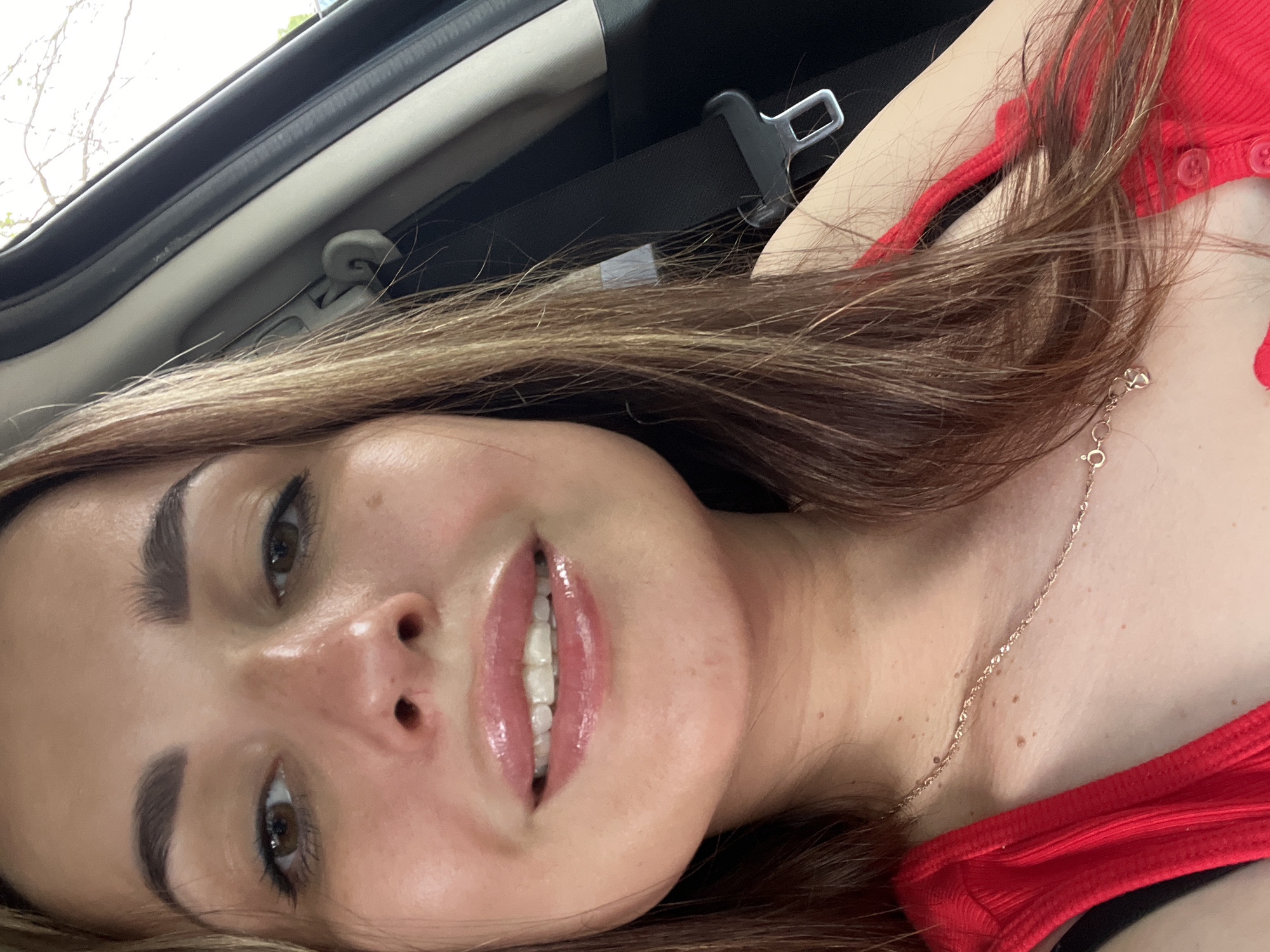 A woman with long, light brown hair and brown eyes is shown from the chest up, in what appears to be the interior of a car. She is wearing a bright red top and a delicate gold necklace with a heart pendant. Her makeup includes dark eyeliner and lip gloss. The background shows a seatbelt and the interior of the car's frame, suggesting she is a passenger or the driver. The lighting is natural and bright, indicating it is daytime.