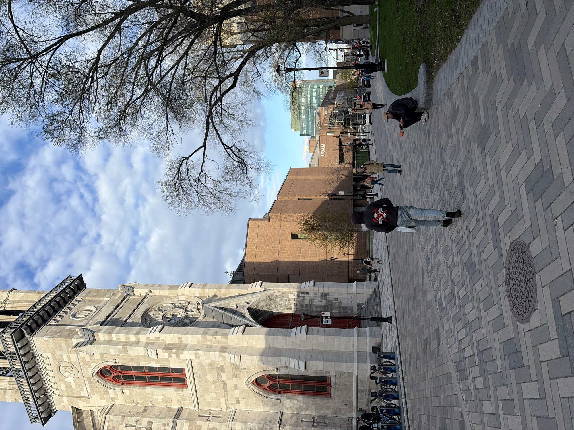This outdoor shot captures a street scene in Montreal, Canada. The image is oriented sideways.
Prominently features the Notre-Dame Basilica on the left, recognizable by its pale stone facade, red-framed windows, and gothic architecture. Adjacent to the Basilica is a modern brick building.
A paved walkway stretches diagonally across the frame. Several people are on the walkway: one person is squatting down, and several others are walking. Streetlights and trees line the walkway.
In the background, a building labeled UQAM is visible, indicating the presence of the Université du Québec à Montréal. Other buildings and the sky are also visible in the background. A bare tree occupies the upper-left corner of the image.
Bikes are parked near the basilica. A manhole cover is visible in the foreground on the pathway.