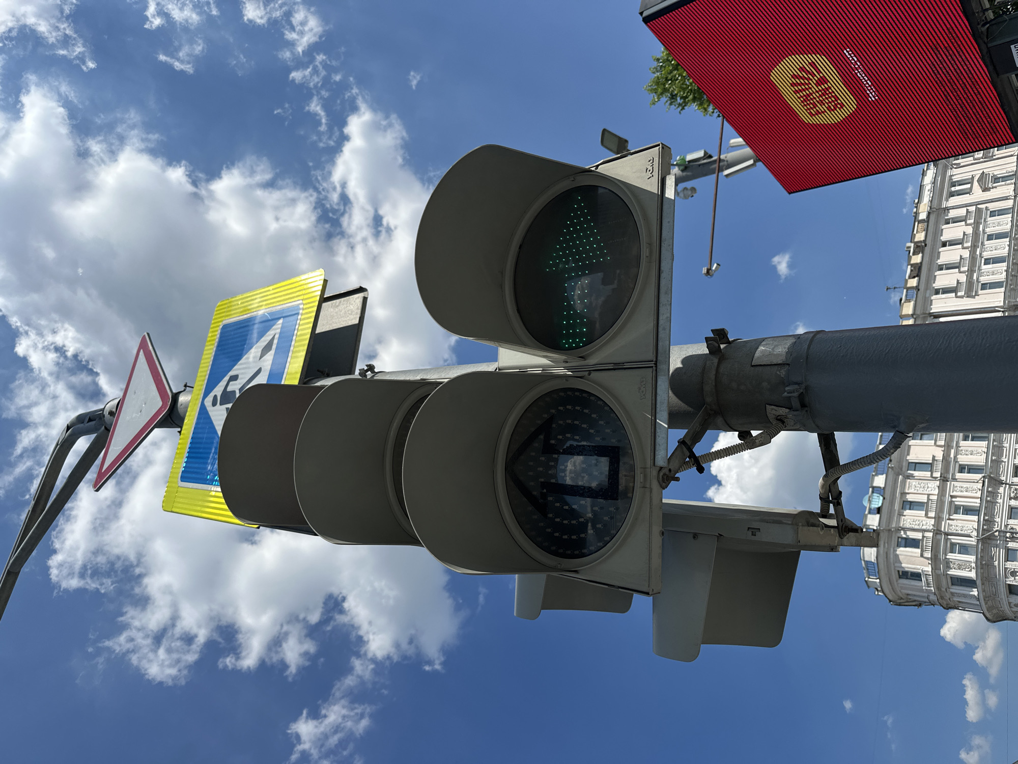 A low-angle, eye-level shot captures a traffic light displaying a green arrow pointing to the right, and a pedestrian crossing sign overhead under a bright blue sky with scattered white clouds. The traffic light is mounted on a pole, which also holds a yield sign and a pedestrian crossing sign. In the background, a building with multiple floors and windows is visible. To the right, a large red digital billboard with yellow lettering is partially visible. The scene appears to be in an urban setting in Moscow, Russia.