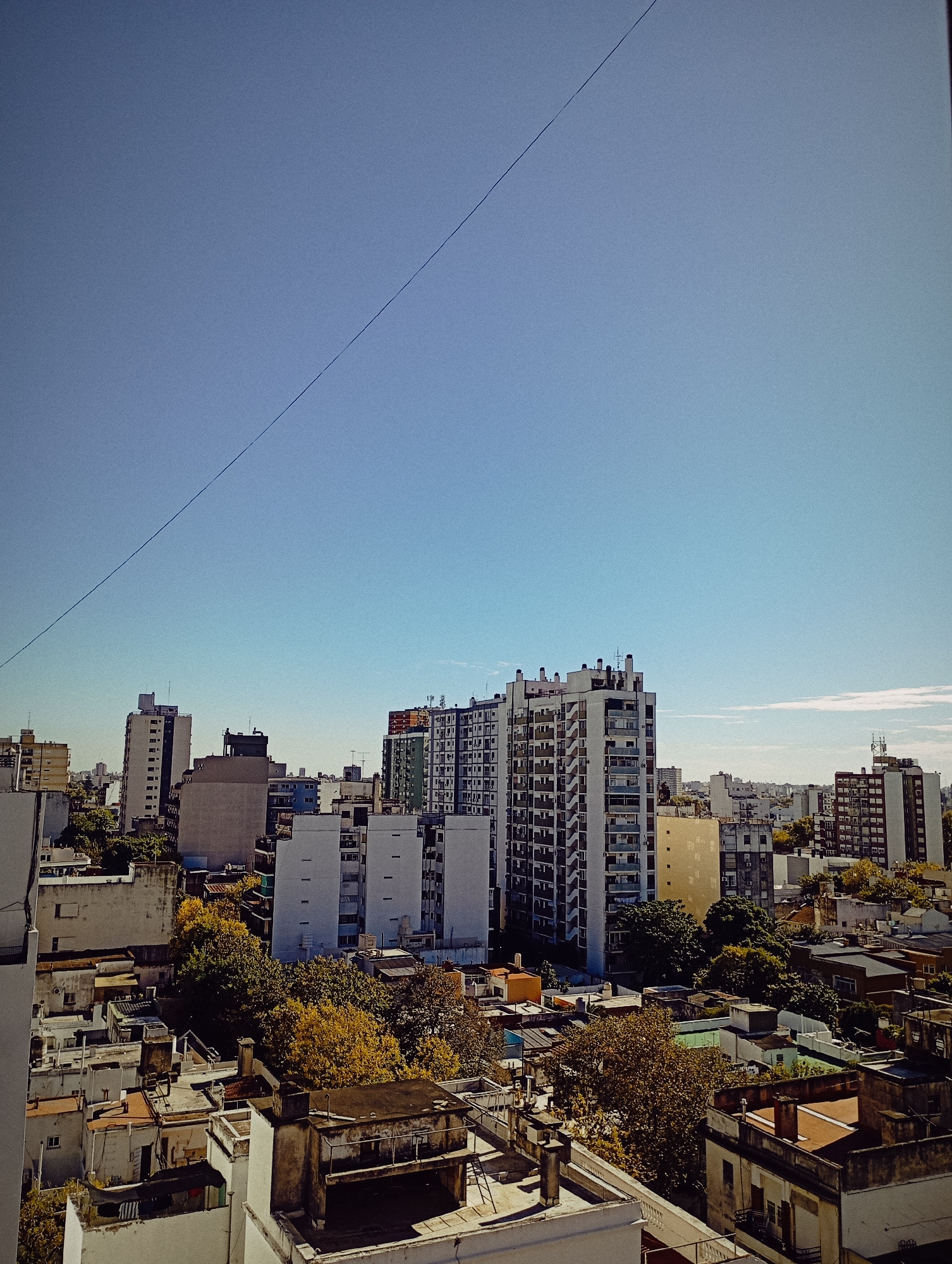 The image presents an urban landscape, predominantly featuring a cityscape of Buenos Aires, Argentina. The foreground is characterized by a dense cluster of rooftops, exhibiting diverse architectural styles and states of maintenance, interspersed with foliage from mature trees, some displaying autumnal hues. This establishes a perspective from an elevated vantage point, such as a high-rise building or rooftop terrace.
The midground is dominated by a series of high-rise apartment buildings, varying in height, color, and design, emblematic of residential architecture common in Buenos Aires. These structures contribute to a relatively uniform skyline, punctuated by the occasional antenna or satellite dish.
The background consists of a clear, blue sky, providing a contrasting backdrop to the urban scene. A thin, dark, diagonal line traverses the sky, suggestive of a power line or cable. The overall setting conveys a typical daytime scene in a densely populated urban environment in Buenos Aires.