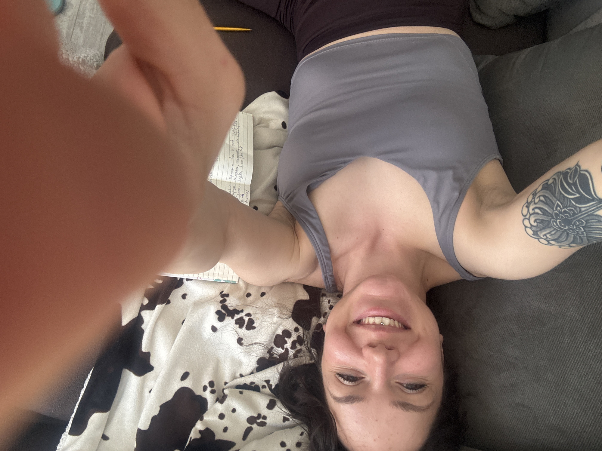 A selfie-style image shows a woman with dark hair lying upside down on a gray couch, smiling at the camera. She is wearing a gray tank top and dark leggings. Her right arm is raised, revealing a tattoo on her upper arm. To her left is a notebook, a cow-print blanket, and a hand, presumably taking the picture. The environment appears to be indoors, possibly a living room. There is no indication of the location of Burgas, Bulgaria, in the image.