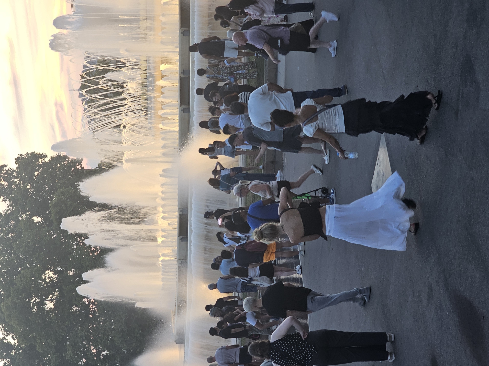 This media file captures a scene featuring a group of people gathered in front of a large fountain display. The setting appears to be outdoors, possibly a public square or park, with trees visible in the background and a paved surface in the foreground. The fountain is actively spraying water, creating plumes of mist and illuminated by soft, warm light, suggesting it is either sunrise or sunset. The sky shows a gradient of colors, with hues of yellow, orange, and pink, reinforcing the idea of dawn or dusk.
The subjects are a diverse group of individuals, all facing the fountain. Many are standing, while some appear to be sitting or kneeling. Their attention is directed towards the water jets, indicating they are observing or enjoying the spectacle. The people are dressed in casual summer clothing. Some are holding drinks or bags. One person is holding a water bottle. There are no visible signs of specific activities like performances or events, but the gathering suggests a shared experience of appreciating the fountain.
Notable details include the gentle lighting that casts a warm glow over the scene, hinting at a pleasant weather day. The overall atmosphere seems relaxed and leisurely. The presence of a large fountain in a public space strongly suggests a tourist or recreational area, and the mention of Barcelona, Spain, places it within that context. The time of day is clearly either early morning or late evening due to the sky's colors and lighting. There is no visible text in the image.
