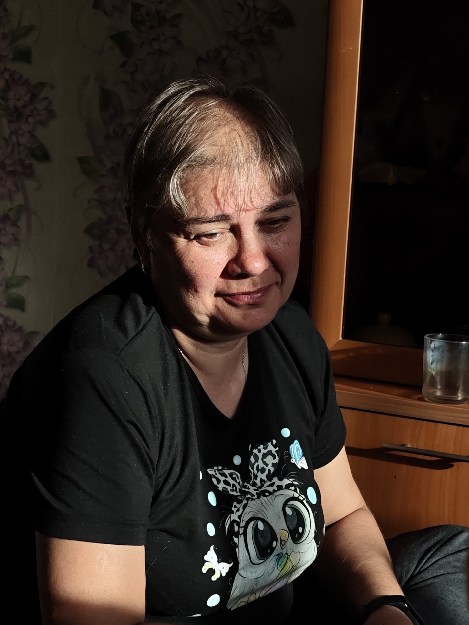 A middle-aged woman with short gray hair is seated indoors, facing slightly downward and to the right. She is wearing a black t-shirt with a cartoon owl graphic. The light is uneven, casting shadows across her face and the patterned wallpaper behind her. To her right, there is a wooden cabinet or dresser with a glass on top. A location is mentioned: Уральское, Russia.