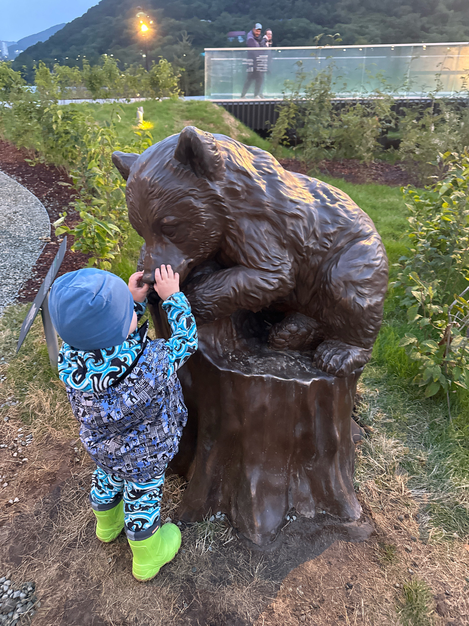 In Petropavlovsk-Kamchatsky, Russia, a young child wearing a blue hat, a blue and black patterned jacket, and bright green boots stands facing a bronze sculpture of a bear on a tree stump. The child is touching the bear's snout with their hands. The sculpture is positioned on a grassy area with dry grass and some dirt. In the background, there are lush green bushes, a walking path with gravel, and further back, a modern pedestrian bridge with railings and lights. Two adults are visible on the bridge. The scene is illuminated by natural light, suggesting it is either early morning or late evening. The overall environment appears to be a park or a landscaped public space with a naturalistic theme.