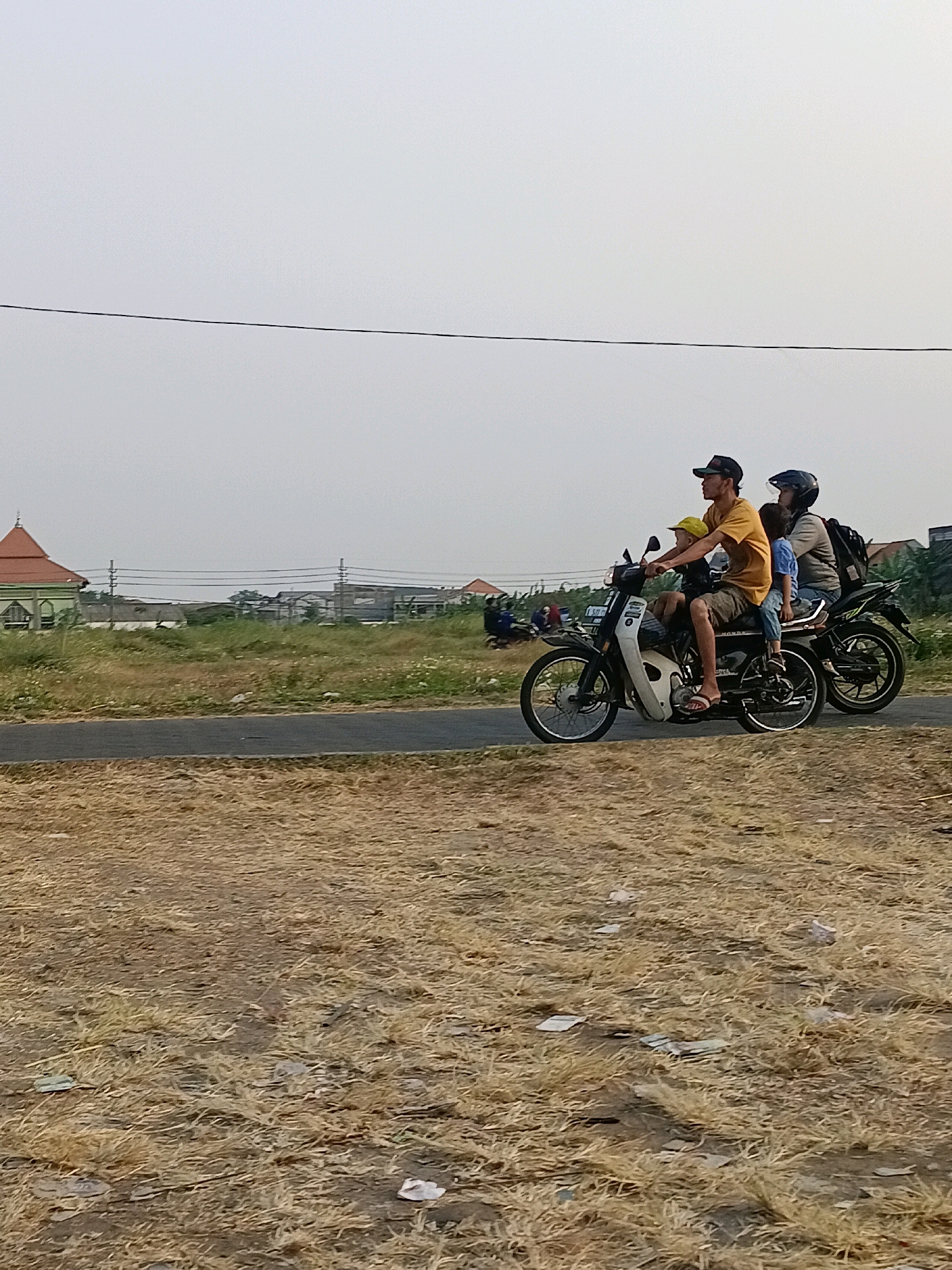 In Tawangsari, Indonesia, a motorcycle carrying three people rides on a paved road. The driver, a man wearing a cap and a yellow t-shirt, sits in front. A young boy in a yellow cap is seated between the driver and the rear passenger. The rear passenger, a person wearing a helmet, is seated behind the driver. They are all moving to the right of the frame. The surrounding environment consists of dry, yellowish grass on the left and right sides of the road, and sparse vegetation further back. In the distance, there are buildings, including a structure with a red roof, and utility poles with power lines. The sky is overcast.