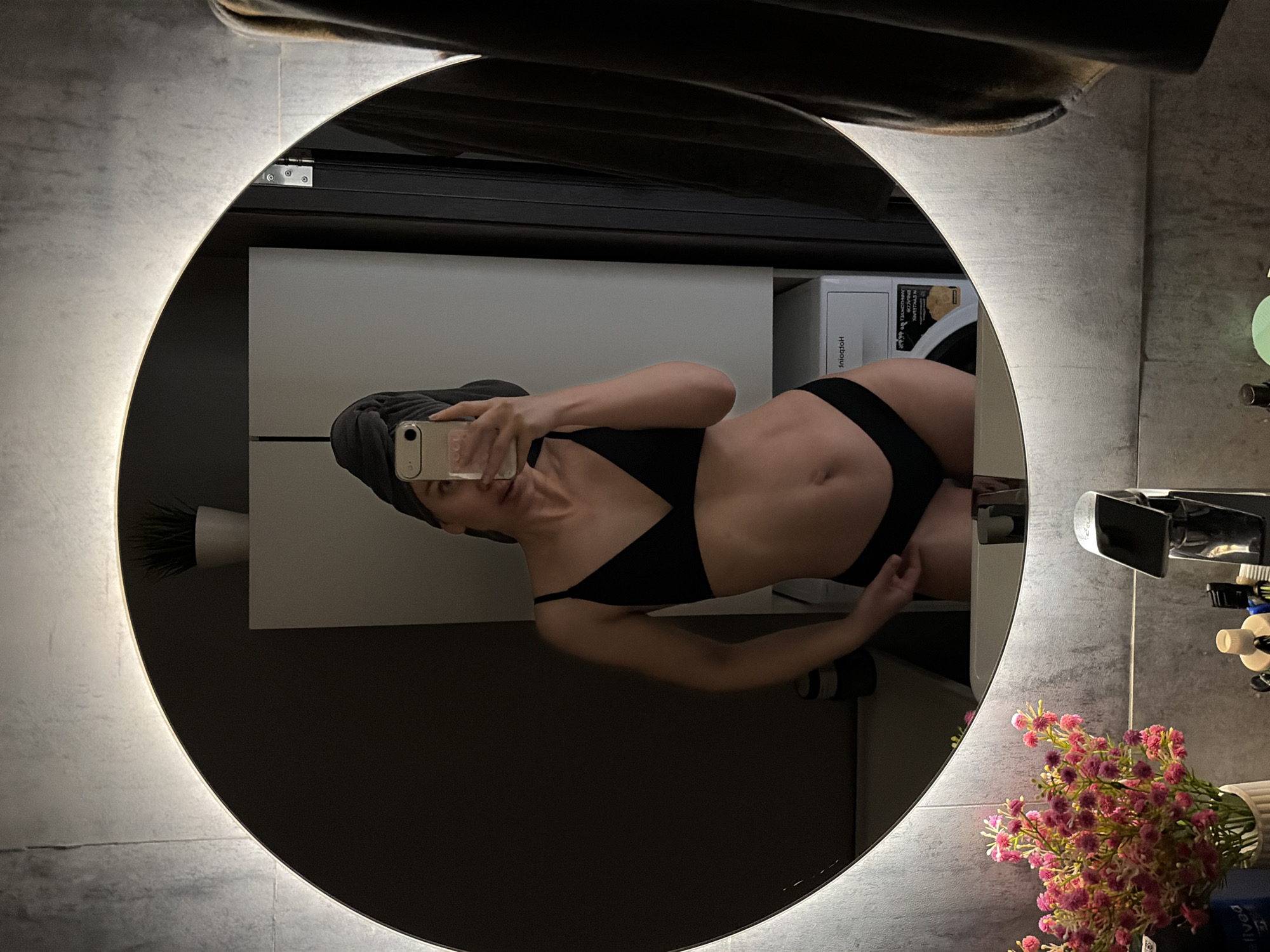 A woman in a black bikini and a towel on her head is taking a selfie in a round, lighted mirror. The mirror reflects a bathroom scene with a white cabinet, a washing machine, flowers in a vase, and bathroom accessories. The location is the city of Chaikovsky, Russia.