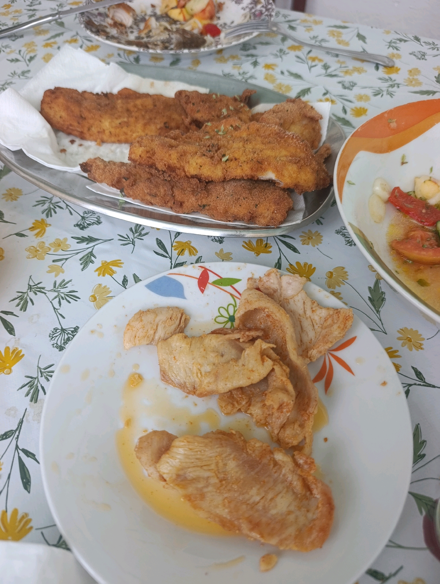 The image depicts a meal on a table covered with a floral tablecloth featuring yellow flowers and green leaves. In the foreground, a white plate contains several pieces of cooked chicken, some of which appear to have been seasoned with paprika or a similar spice, and are resting in a light yellow sauce. Behind this plate, a metallic tray holds several pieces of breaded and fried food, possibly fish or chicken, served on white paper towels. To the right of the fried food, a white bowl with an orange rim contains a salad or appetizer with chopped tomatoes, white pieces (possibly hard-boiled eggs or cheese), and a light dressing. In the upper background, another plate with remnants of food and a fork is visible. The lighting suggests an indoor setting, possibly a restaurant or dining room. The presence of various dishes suggests a meal in progress or recently consumed.
Based on the visual cues, specifically the floral tablecloth and the style of the dishes, this scene is highly characteristic of a traditional Portuguese restaurant, particularly in the region of Setúbal, Portugal. Such tablecloths are common in local eateries, and the cuisine presented, featuring fried fish and simple salads, aligns with regional culinary traditions. Setúbal is known for its excellent seafood and traditional Portuguese cuisine, making it a plausible location for this dining scene.