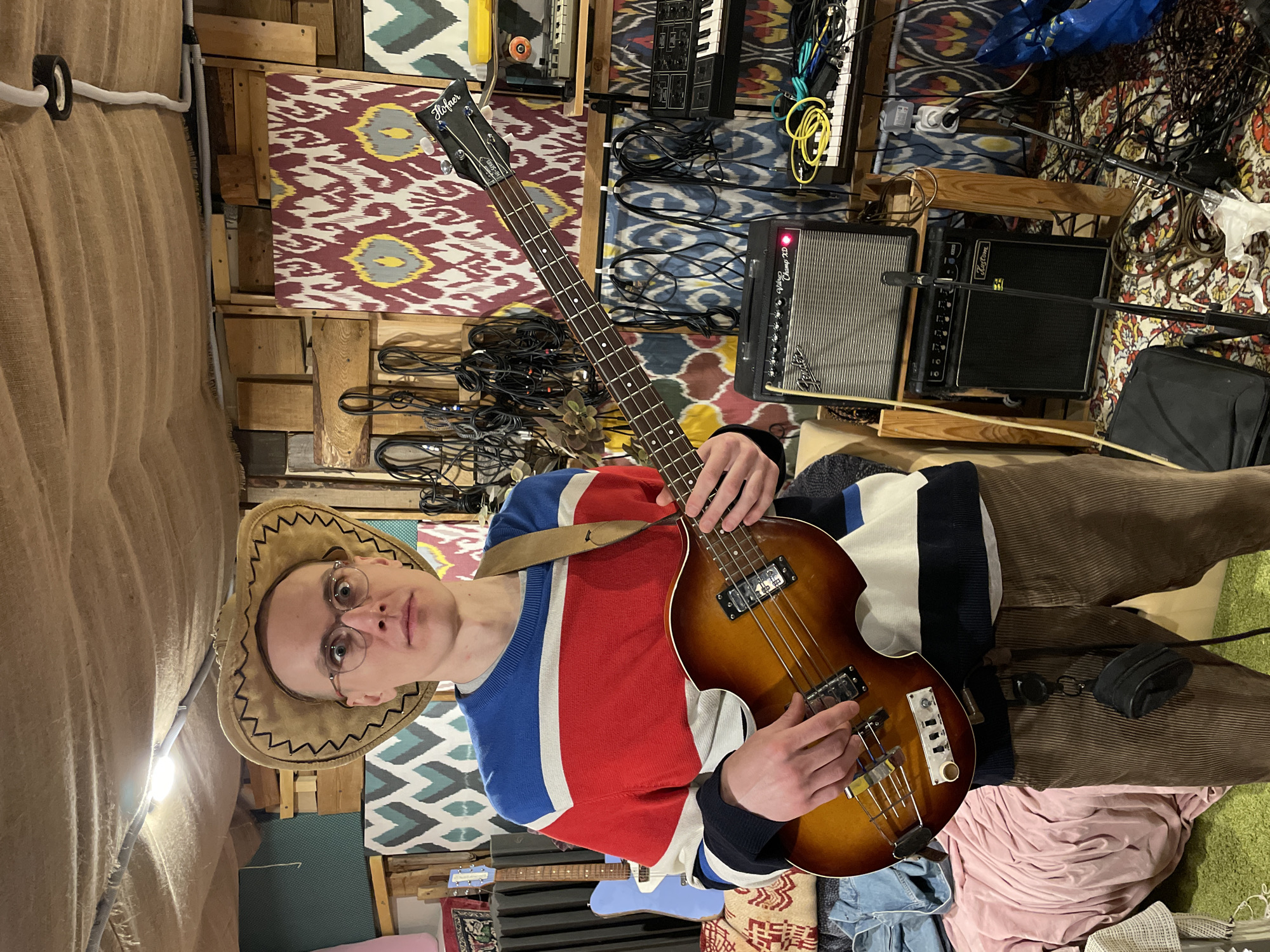 A young man with glasses and a cowboy hat stands in a room holding a Hofner bass guitar. He wears a red, white, and blue sweater and brown corduroy pants. Behind him is a wall decorated with wooden crates and colorful patterned fabric. Wires and musical equipment are visible in the background, including keyboards and amplifiers. The location is not provided in the image.