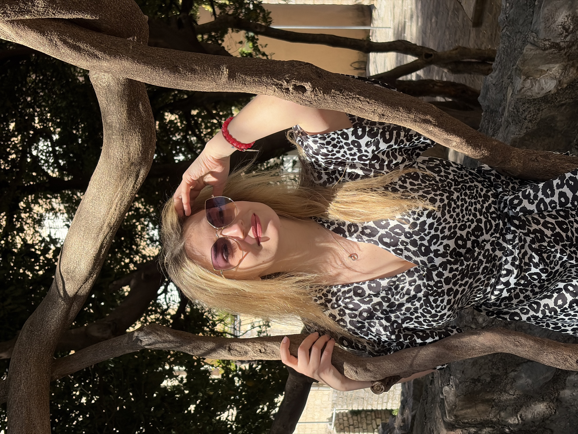 A woman with blonde hair and sunglasses is posing outdoors in what appears to be the Old Town of Budva, Montenegro. She is wearing a black and white animal-print dress and a red bracelet on her left wrist. She has one hand on her head and another holding onto a tree branch. The tree has thick, brown branches. In the background, there are stone walls and more trees. The photo is taken at an angle, with the woman tilted to the side.