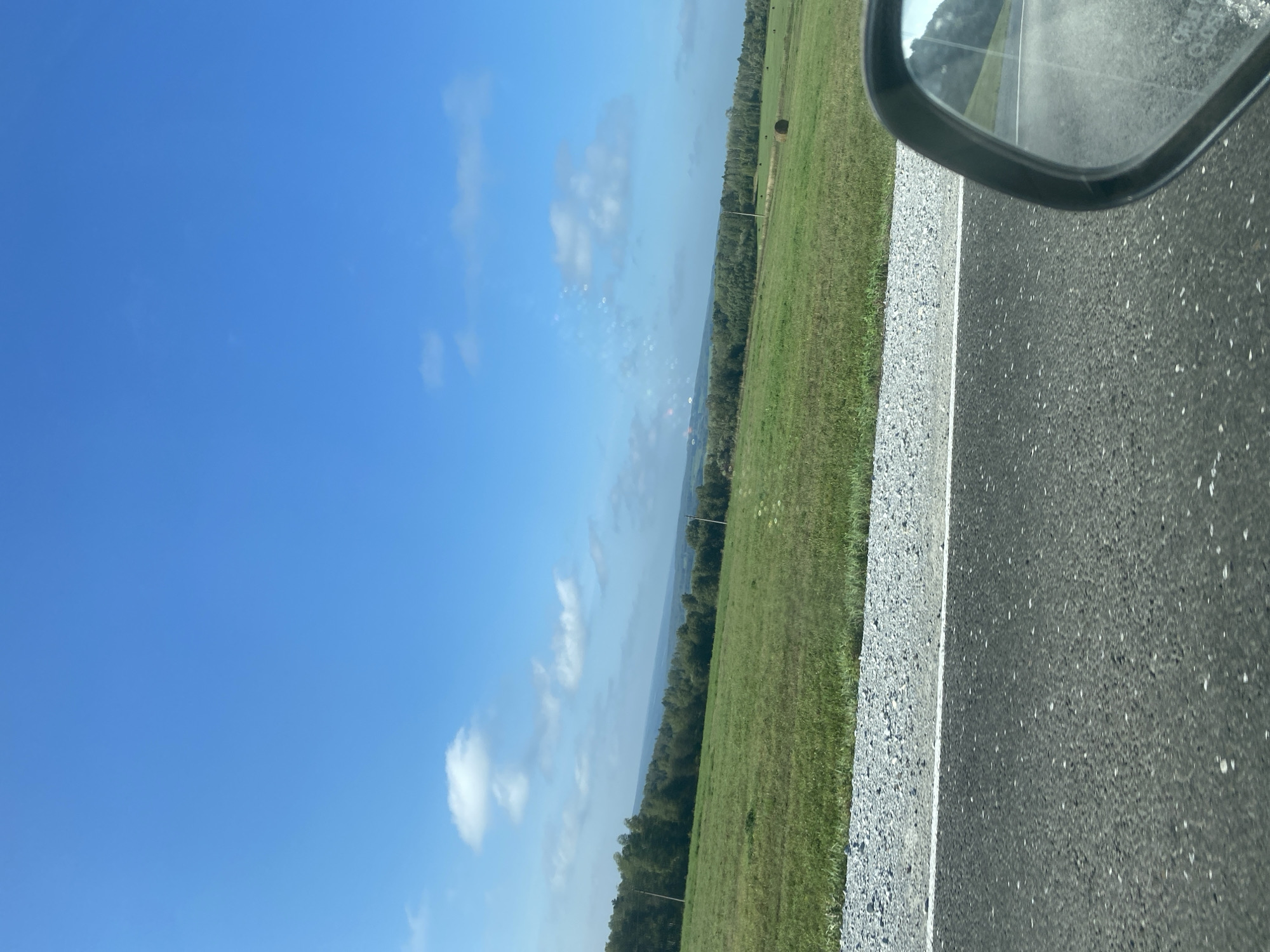 The image depicts a rural landscape under a bright blue sky with scattered white clouds. In the foreground, an asphalt road is visible, bordered by a gravel shoulder. To the left of the road, a vast green field stretches towards a line of trees in the distance. Several round hay bales are scattered across the field. A car's side mirror is visible in the lower right corner, reflecting a portion of the road and the sky. There are no people or specific structures indicative of Podonino, Russia, present in the image.
