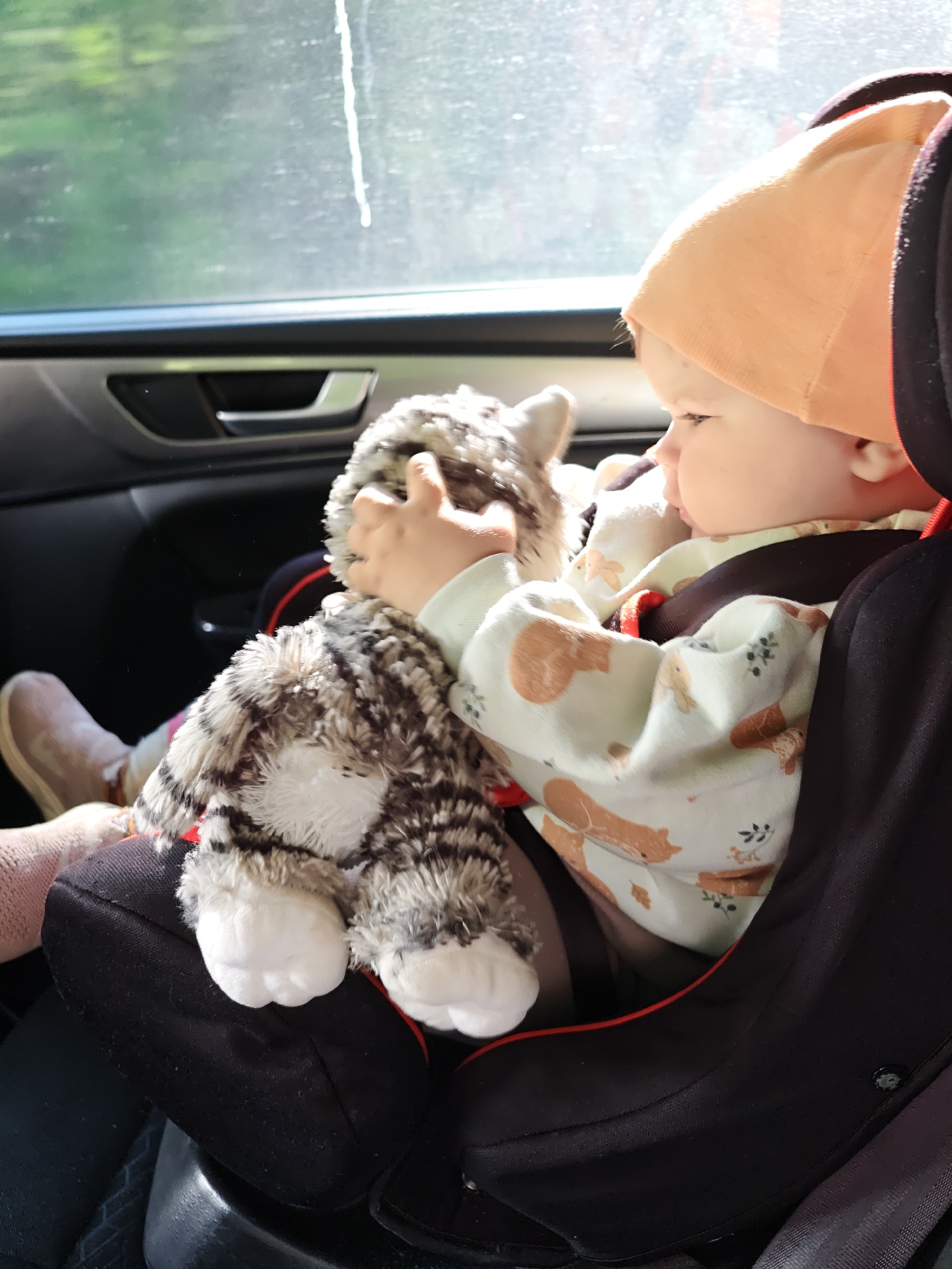 A baby wearing a peach-colored hat and a light-colored shirt with a print is seated in a black car seat within a vehicle. The baby is holding a stuffed animal resembling a cat. The scene is lit by sunlight coming through the car window. The location, Oralki, Russia, is not evident in the image.
