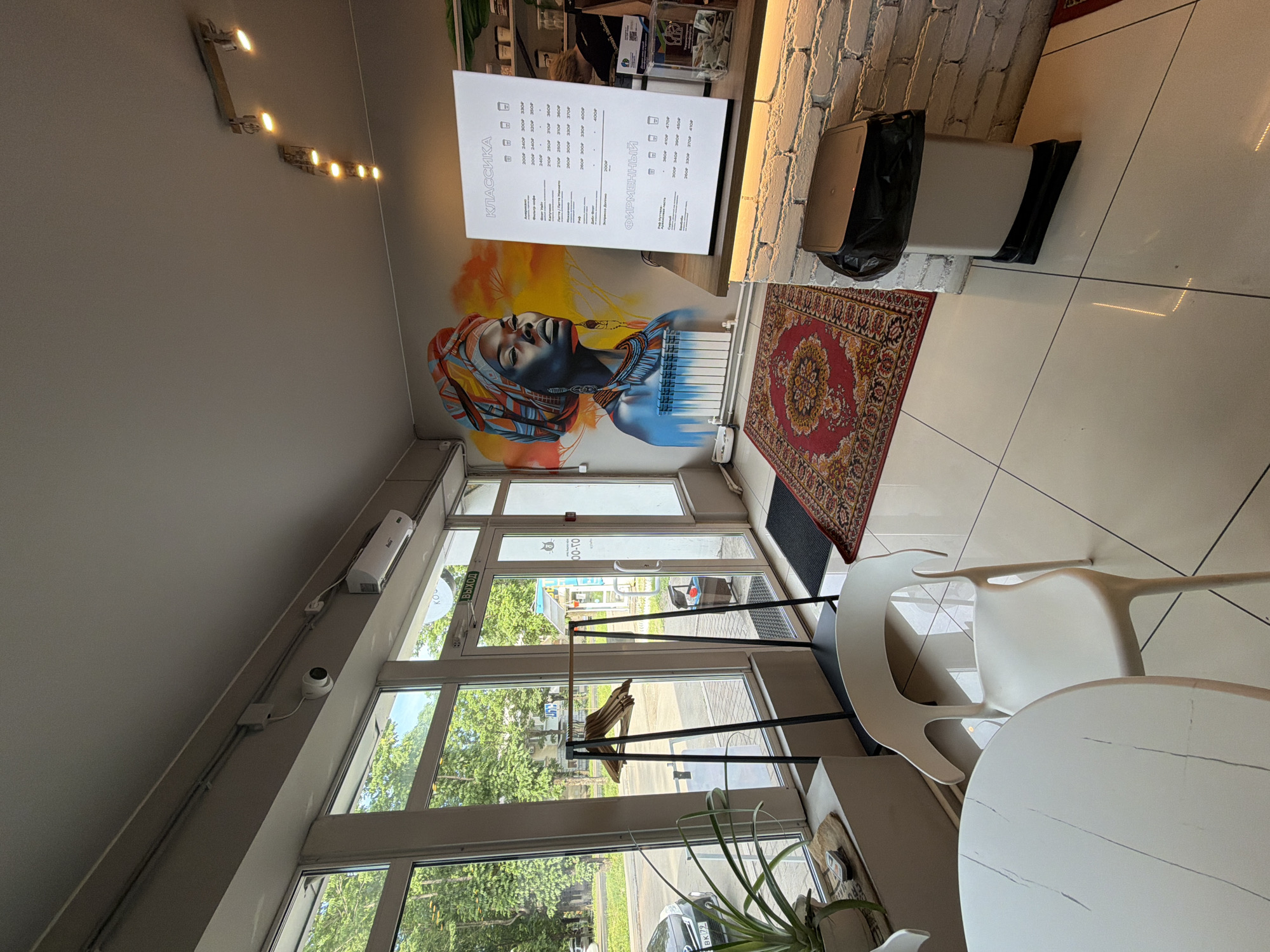 The image shows the interior of a cafe in Birobidzhan, Russia. The cafe features a large mural of a woman with a colorful headwrap on one wall. A menu is displayed on a stand on the counter. There are white tables and chairs in the foreground, and a rack with hangers by the window. The floor is tiled. A trash can is visible near the counter. The overall impression is of a modern and stylish cafe.