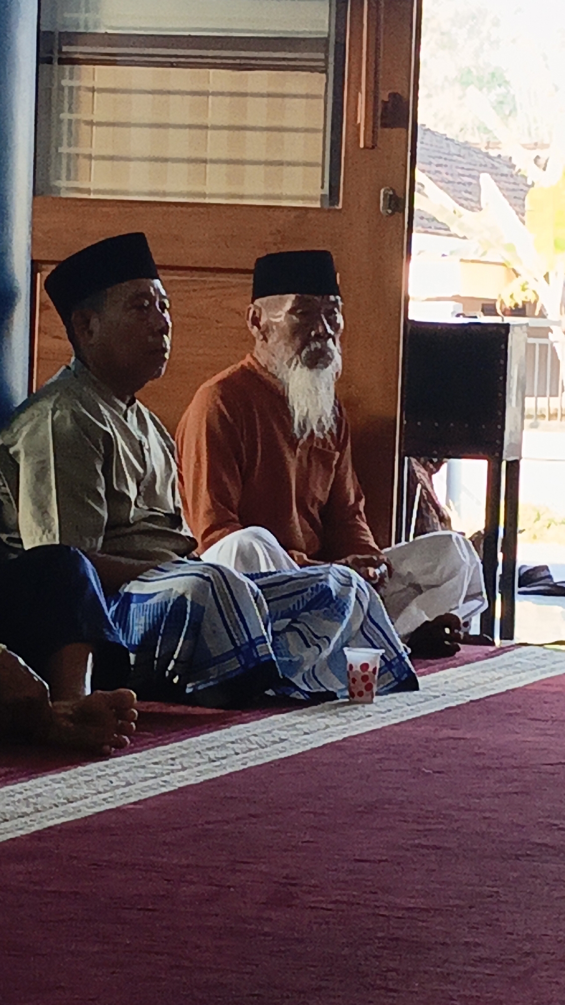 Two men in traditional Islamic attire are sitting on the floor in what appears to be a prayer or gathering space. The man on the left wears a light-colored tunic and a black cap. The man on the right has a long white beard, wears an orange tunic, and a black cap. Both are seated cross-legged on a carpeted floor, with the man on the left wearing a sarong. In front of them, on the floor, is a red and white striped carpet. A small, clear plastic cup with red polka dots is visible near the man on the right. The background shows a wooden doorway and blinds on a window, suggesting an indoor setting with natural light. This scene is located in Srono, Indonesia.