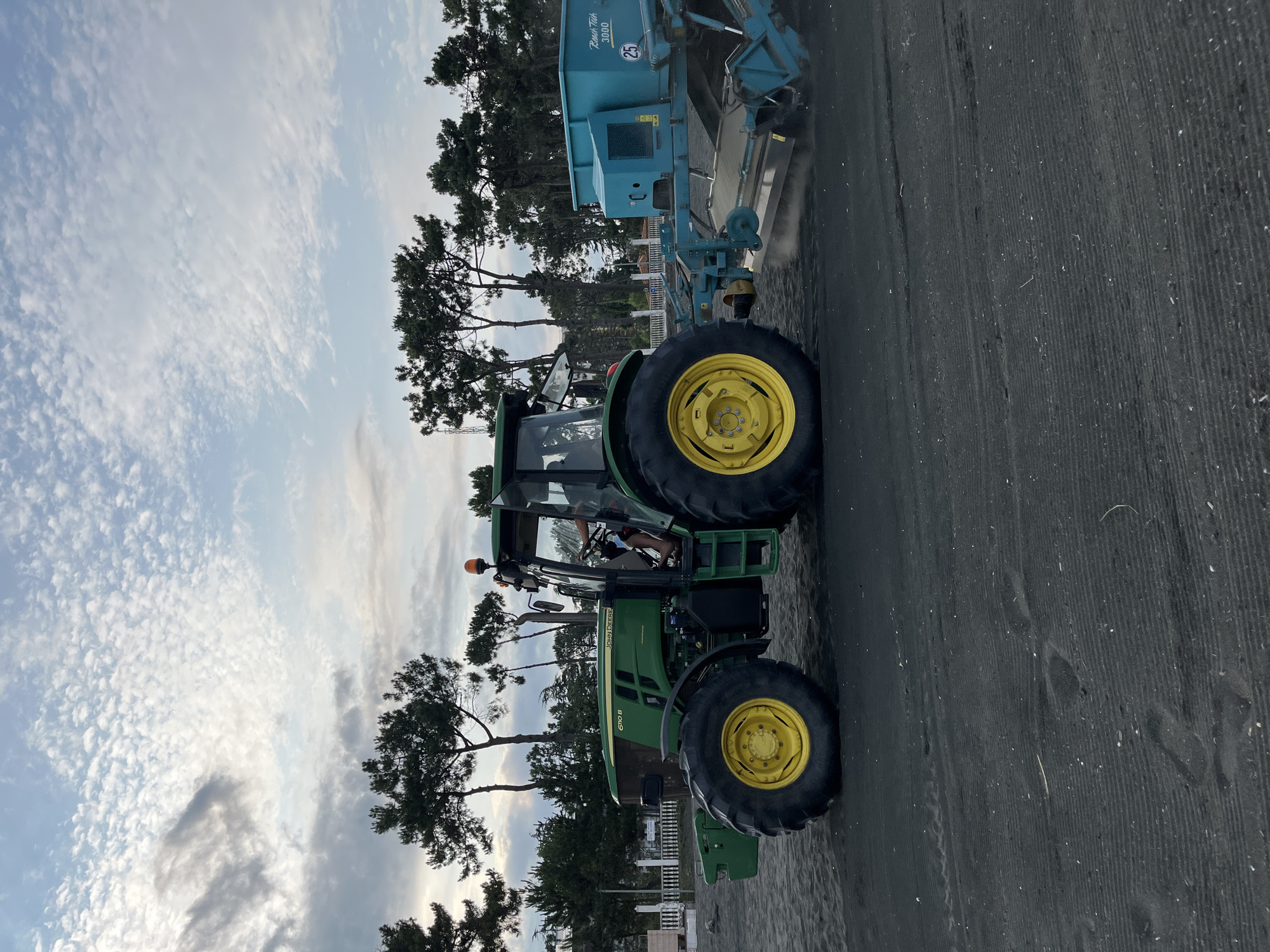 In Ureki, Georgia, a green John Deere tractor is working on a black sand beach. The tractor is pulling a turquoise-colored sand grooming attachment. The attachment appears to be in operation, as evidenced by the dust or sand being kicked up by the rotating tines at the rear. The tractor has large, yellow-rimmed wheels and the model designation "6110 B" is visible on the side. A person is visible in the driver's seat, operating the tractor. The background features a line of tall, slender pine trees against a cloudy sky. A white fence and some buildings are also visible behind the trees. The sand is dark and textured, with some small shells and debris scattered on its surface. The overall scene depicts agricultural or maintenance work being performed on a beach environment.