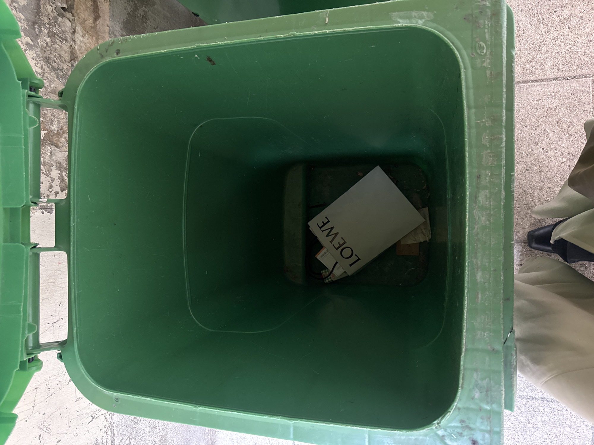 The image is a top-down shot of an open green trash can. Inside the can, there's a Loewe branded item, likely a piece of paper or packaging. The trash can is situated on a light grey tiled surface. To the right of the can, part of a person's leg is visible, clad in beige fabric and a black shoe. The location is identified as Paris, France, presumably within an urban environment.