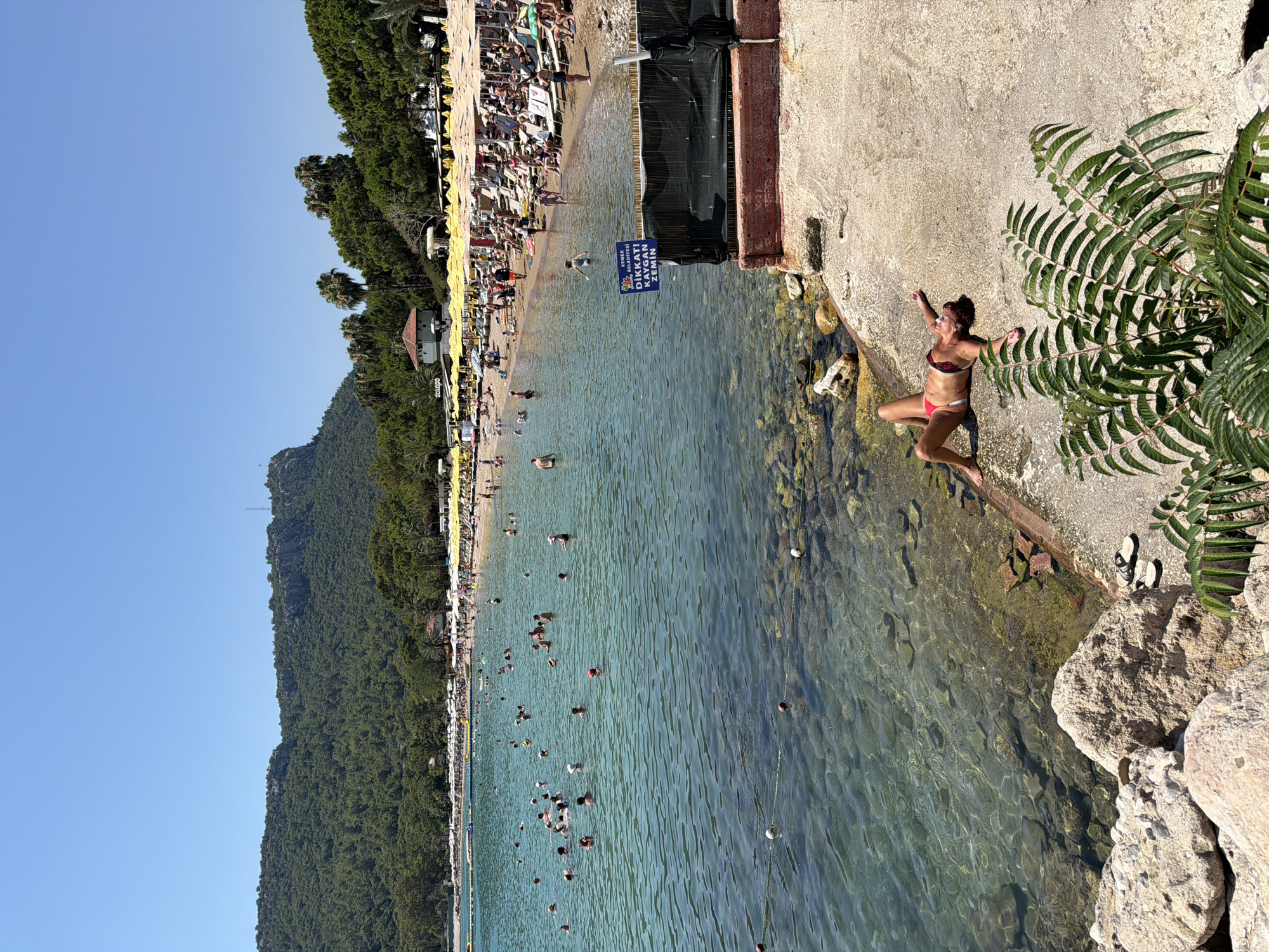 The image depicts a crowded beach scene in Kemer, Turkey, on a clear, sunny day. A woman in a red bikini lies on a concrete ledge near the water, facing upwards and appearing to be sunbathing. The water is clear and turquoise, with numerous people swimming and wading. The beach is lined with rows of yellow umbrellas and lounge chairs, indicating a resort or public beach area. In the background, a lush, green forested mountain dominates the landscape, with a prominent rock formation at its peak and a tower. A sign with Turkish writing, likely a warning, is visible near the foreground. The overall impression is one of a popular summer vacation destination.