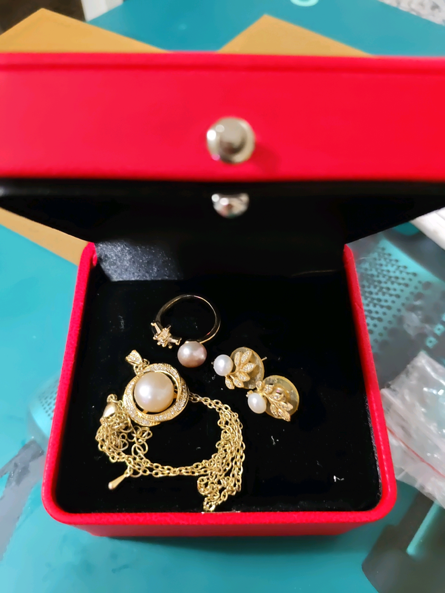 The image shows a jewelry box containing a gold necklace with a pearl pendant, a pearl ring, and a pair of pearl earrings. The jewelry is placed on a black velvet lining inside a red box. The background is a blurred turquoise surface. The image does not provide any information about the location in the city of Rostov-on-Don, Russia, nor does it depict any people, actions, or interactions.