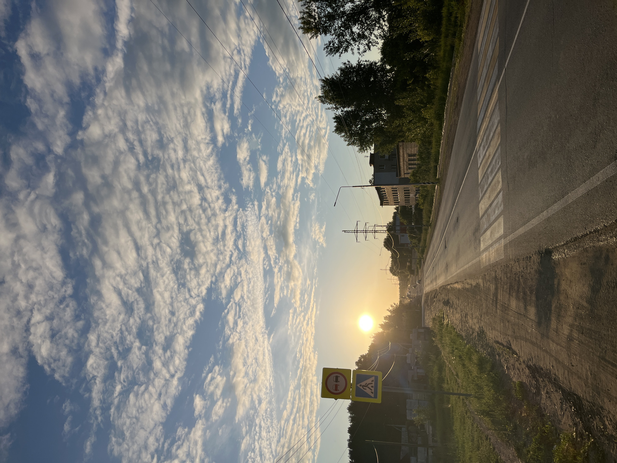 The image depicts a semi-urban or suburban street scene at either sunrise or sunset, characterized by warm, golden light and long shadows. The sky features scattered altocumulus or stratocumulus clouds against a blue backdrop.
In the foreground and left side, a dirt and grass shoulder parallels an asphalt road. A pole displays two yellow-backed road signs: a circular "No Overtaking" sign with two red cars crossed out, mounted above a square "Pedestrian Crossing" warning sign depicting a walking figure. Behind these signs, stacks of concrete blocks or building materials are visible, followed by a dense line of trees along the horizon.
The asphalt road extends into the distance, illuminated by the low, bright sun positioned on the left side of the frame, near the horizon. A pedestrian zebra crossing, marked with faded white and yellow stripes, is visible on the road surface in the mid-ground. White solid lines delineate the road's edges. Faint outlines of vehicles can be seen in the distance, heading towards the sun's glare.
On the right side of the road, a strip of grass and trees gives way to architectural structures. A multi-story, light-colored building with numerous windows, possibly residential or administrative, stands prominently, accompanied by a smaller, older-looking brick building. A black streetlight pole with a single lamp is visible near these buildings. Multiple utility poles and power lines crisscross the sky, and large metal lattice power pylons are discernible in the farther distance.
No individuals or animals are visible within the frame. The road signage is consistent with standards used in the Russian Federation. However, specific visual landmarks within the image are insufficient to forensically pinpoint an exact location within the city of Chaikovsky, Russia.