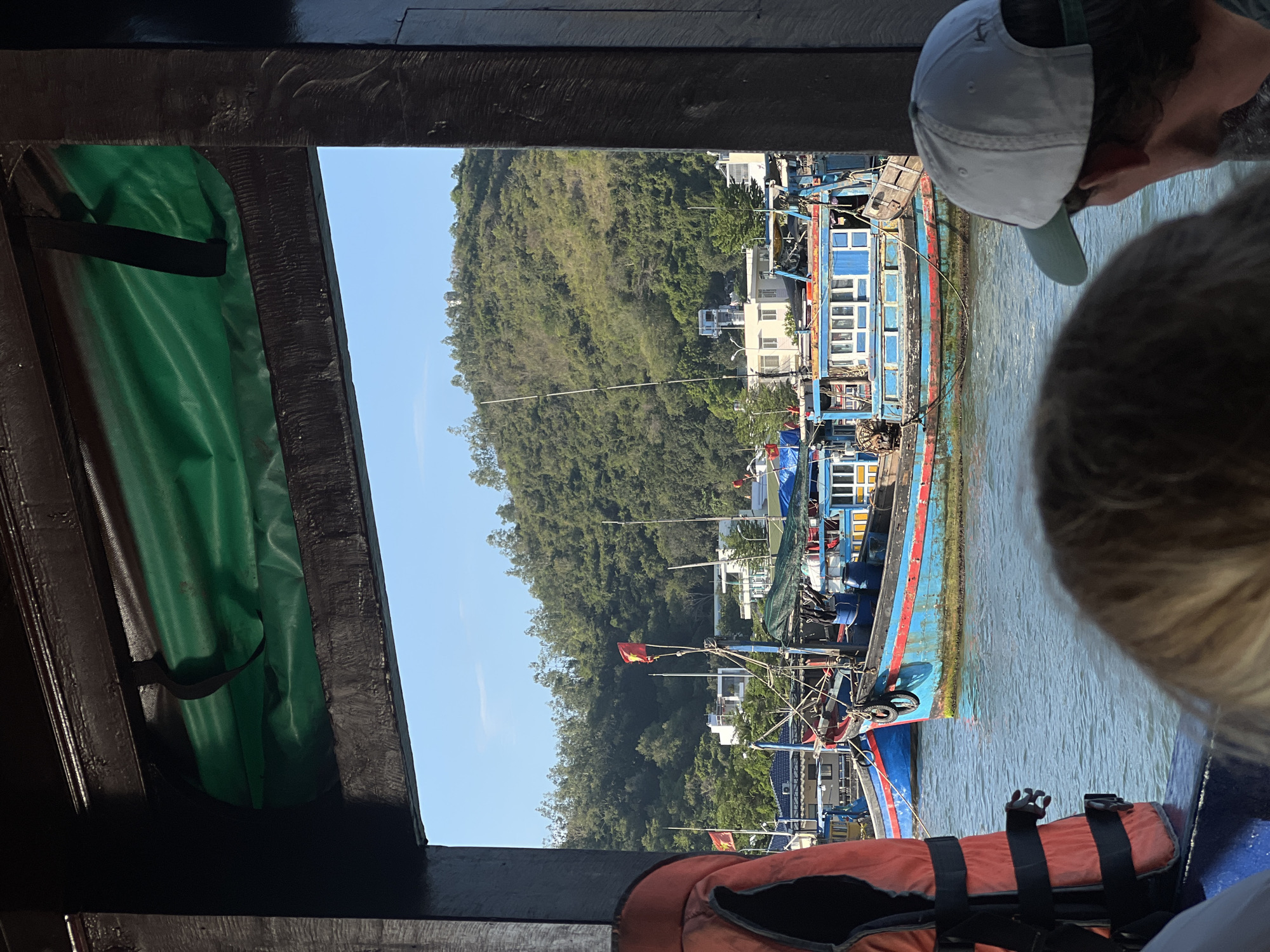 A medium shot taken from inside a boat shows several other boats in a harbor in Nam Nha Trang Ward, Vietnam. One person wears a light gray baseball cap. A life vest sits on the floor of the boat from which the picture is taken. A lush green hillside rises behind the docked boats. Many of the boats appear to have faded blue paint.