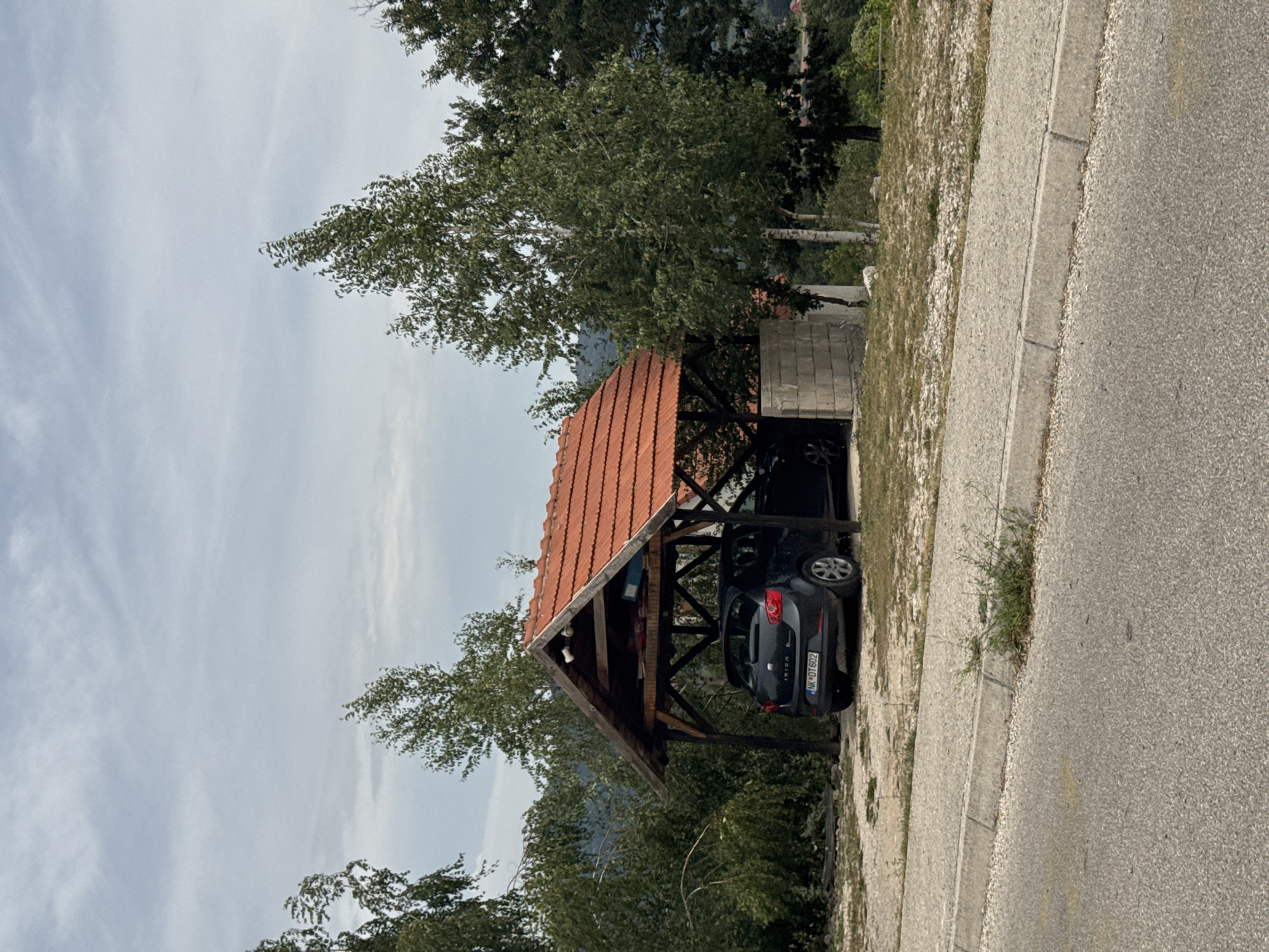 In the city of Vidrovan, Montenegro, a dark-colored hatchback car is parked under a wooden carport with a red-tiled roof. The carport is supported by wooden beams and has a partial wall made of cinder blocks on one side. The car's license plate reads "NK DT 602". The surrounding environment includes several trees with green foliage, a cloudy sky, and a paved road with a curb in the foreground. The ground around the carport is covered in gravel and dry grass.