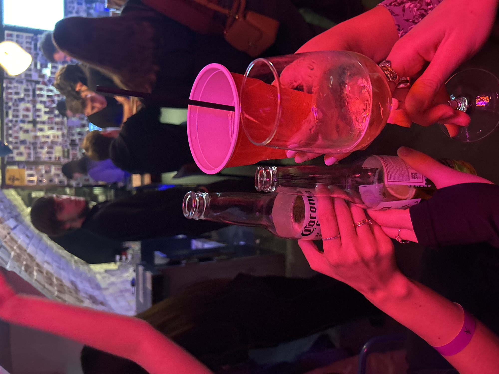 In a dimly lit bar-like environment, multiple people are gathered, illuminated by red and pink lighting. Foreground elements include an overhead view of three sets of hands holding drinks. The left-most hands hold a red plastic cup with a black straw. Next to it, a pair of hands grasps a large, clear wine glass containing a reddish beverage. The right-most hands hold two brown glass bottles of Corona beer. Other blurry people are visible in the background, along with a wall decorated with photos. The exact location in Krasnoyarsk, Russia is not identifiable from the image.