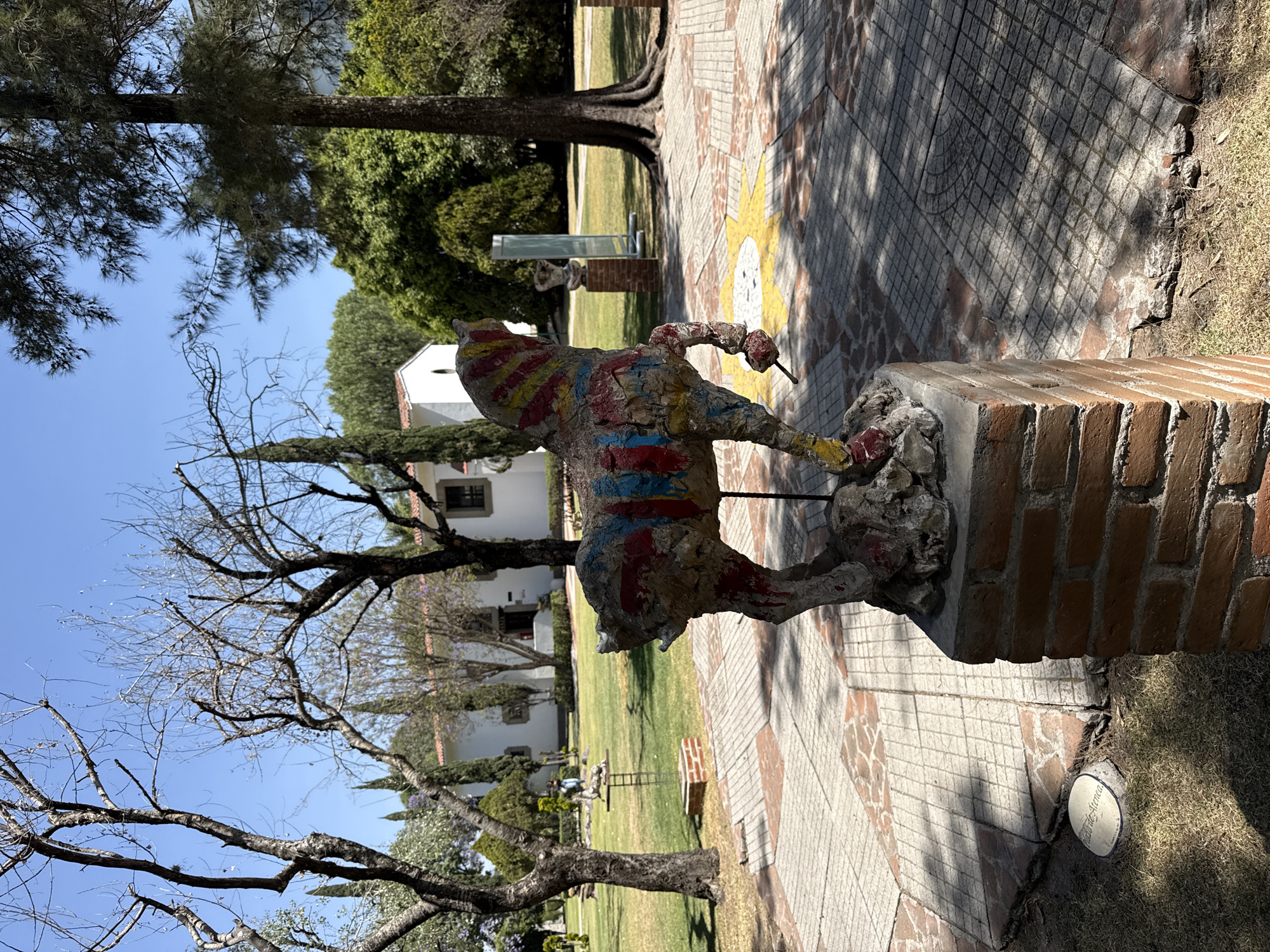 An outdoor sculpture garden in San Andrés Cholula, Mexico, is depicted under a clear blue sky. A multi-colored abstract horse sculpture, predominantly grey with irregular red, blue, and yellow painted stripes, stands on a reddish-brown brick pedestal in the foreground. The sculpture faces left, with its front right leg raised. The ground features a patterned paved walkway composed of grey square tiles and reddish-brown sections, one of which displays a yellow sun motif. Patches of green and dry grass flank the paved areas. A small, round white ground marker with partial dark text is visible on the grass near the main sculpture's pedestal. In the mid-ground, a white stucco building with a reddish-tiled roof is present, surrounded by various trees, some bare-branched and others fully foliated, including tall cypress-like trees and a large pine tree on the right. Multiple other sculptures, including a bust on a brick pedestal and a tall, reflective structure, are dispersed across the grassy areas. A single individual is observed walking in the distant background near the white building.