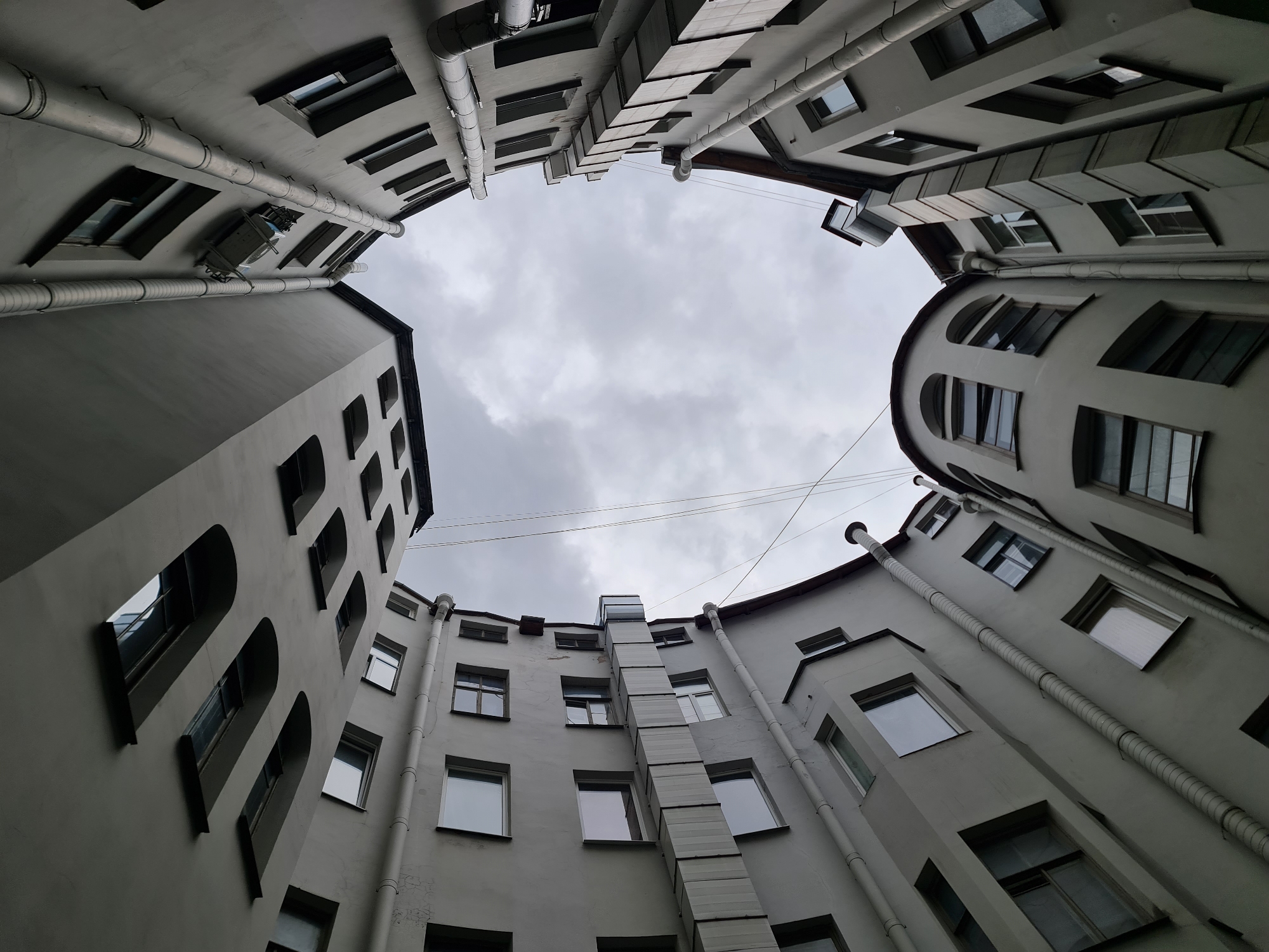 This is a low-angle, wide shot of a courtyard in Saint Petersburg, Russia, looking up at the surrounding buildings. The buildings are light gray with numerous windows and external pipes. The sky is overcast and gray. The architecture suggests a typical pre-revolution building, possibly in the historic center of the city.