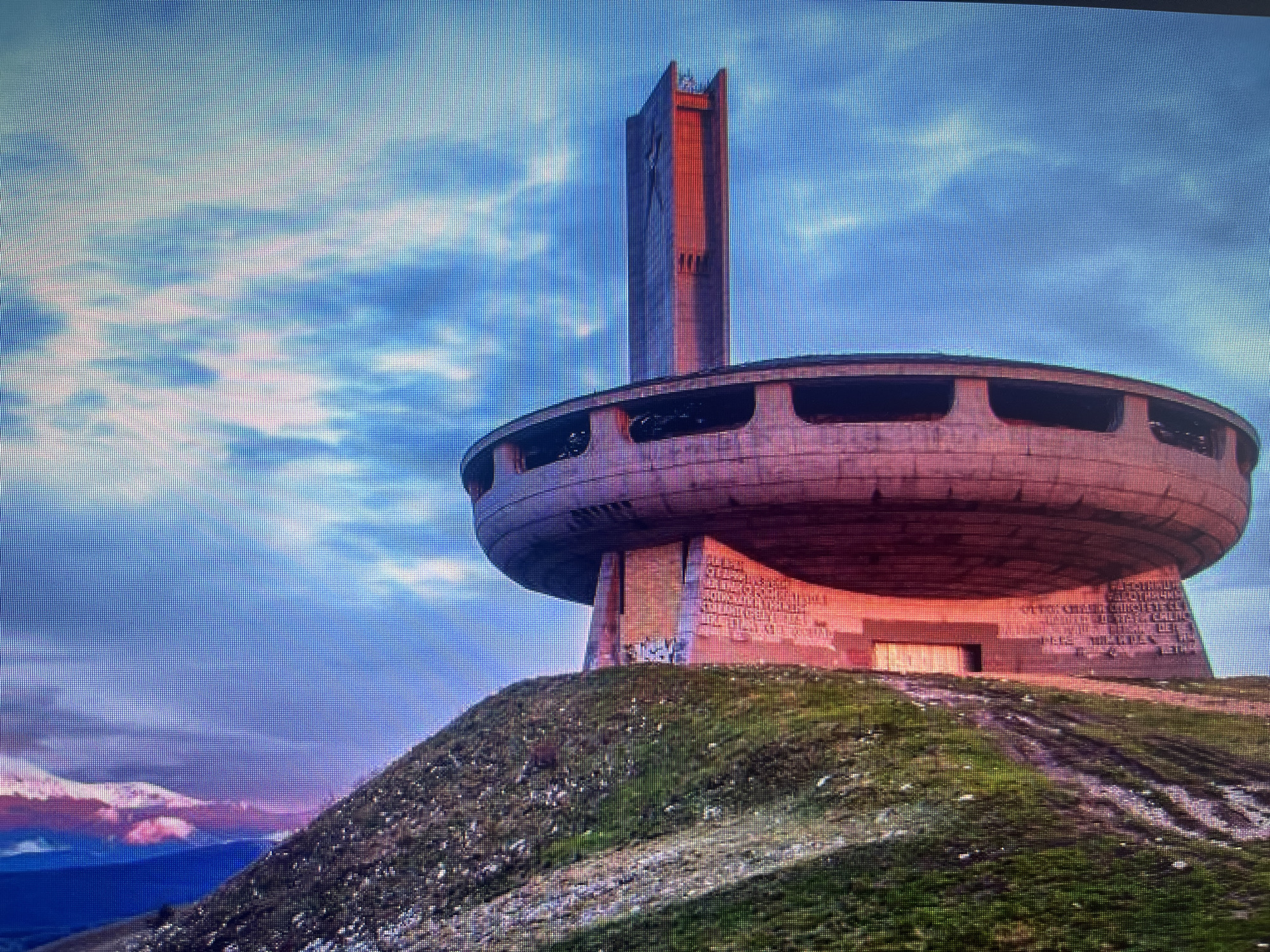 An eye-level, wide shot captures the imposing Buzludzha Monument, a brutalist concrete structure, atop a grassy mountain peak under a dramatic sky. The monument consists of a large, circular, saucer-shaped building with numerous rectangular window openings, connected to a tall, rectangular tower that rises above it. The tower features vertical design elements and what appears to be a star symbol near its apex.
The scene is bathed in the warm, reddish-orange light of either sunrise or sunset, highlighting sections of the concrete facade, particularly on the tower and the underside of the circular structure. The sky above is partly cloudy, displaying shades of blue, white, and purple, with some distant clouds over mountains also catching purplish hues. The monument itself appears weathered and somewhat dilapidated, with some graffiti visible on its base, suggesting an abandoned state.
The monument sits on a verdant hill, with worn dirt tracks leading up to its base. On the concrete facade supporting the circular building, Cyrillic inscriptions are carved, though largely unreadable due to distance and resolution. No people are visible, emphasizing the monument's isolated and monumental scale in the landscape. The overall mood is one of stark grandeur and historical weight.