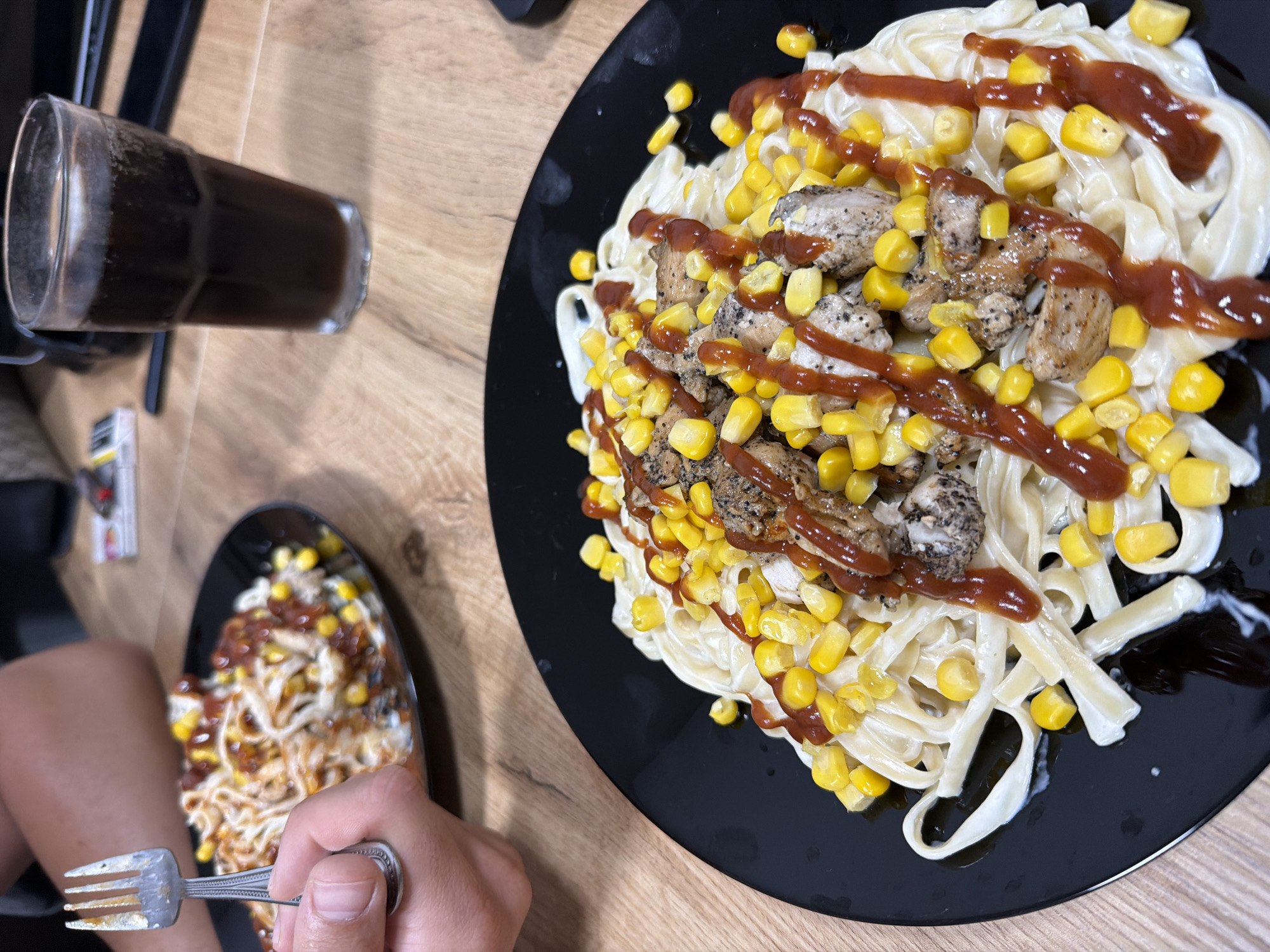 The image depicts a close-up view of a black plate filled with fettuccine pasta, topped with pieces of seasoned chicken, corn kernels, and a drizzle of ketchup or a similar red sauce. In the background, another plate of pasta with corn and sauce is visible, along with a hand holding a fork. A glass of dark beverage, possibly soda or iced tea, and a pack of cigarettes are also present on a wooden table. The setting appears to be a casual dining environment. There is no information in the image to determine the location in the city of Plovdiv, Bulgaria.