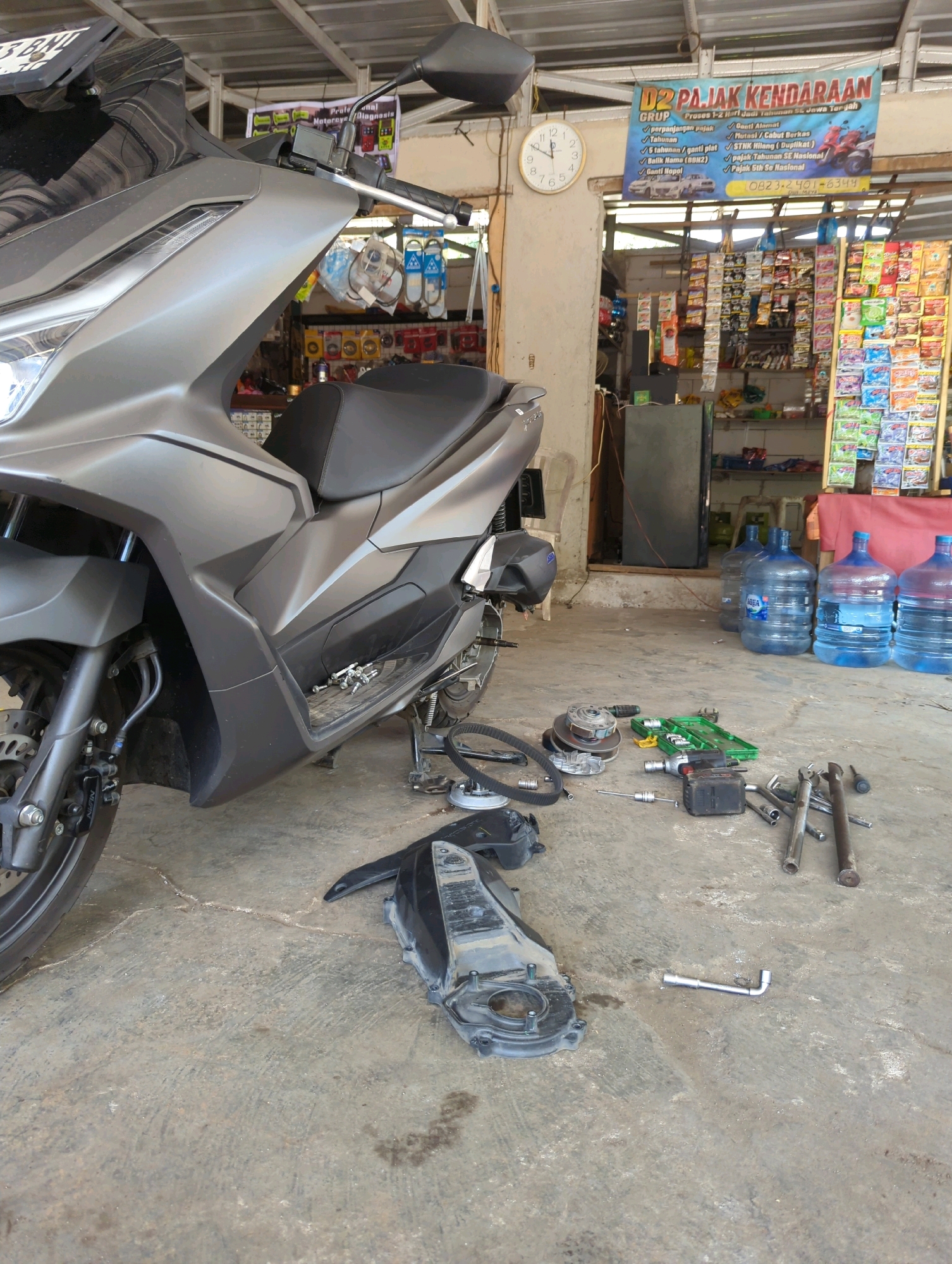 This is a medium shot, eye-level, full-color, outdoor photograph of a motorcycle repair area in the city of Senggrong, Indonesia. The focus is on a silver scooter, partially disassembled, with various tools and parts scattered on the concrete floor. A large banner for "D2 PAJAK KENDARAAN" (Vehicle Tax Services) hangs on the wall behind the scooter, with contact information and a list of services offered. A clock on the wall indicates the time is approximately 9:50. Several large water jugs are visible to the right of the scooter, along with shelves stocked with various goods. The overall scene suggests a busy, utilitarian workspace.