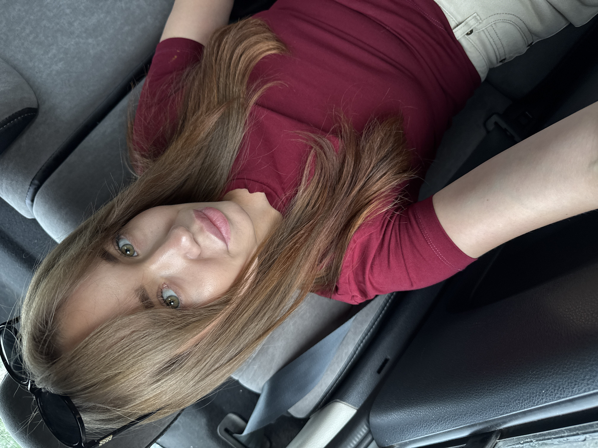 In a dimly lit interior, likely the back seat of a car, a young woman with long, light brown hair and green eyes gazes at the camera. Sunglasses are perched on her head, and she wears a maroon t-shirt and off-white shorts. The background consists of car seats and a seatbelt, suggesting a journey. The scene is devoid of any specific landmarks or indicators that would place it in Chaikovsky, Russia, or any other particular city.