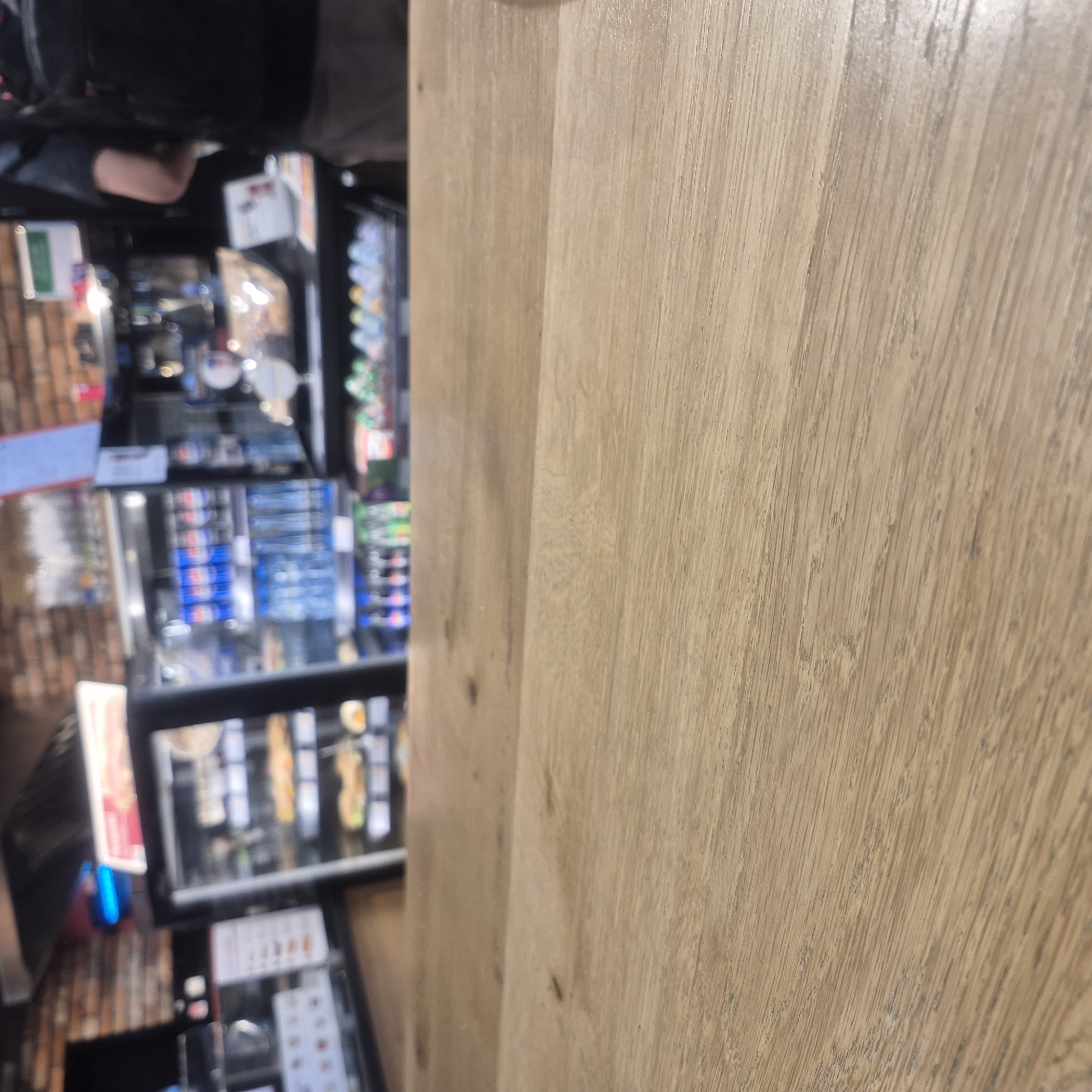 The image displays a vertical shot of what appears to be an interior setting, possibly a convenience store or a small shop. The foreground is dominated by a light-colored wooden surface, likely a counter or shelf, with visible wood grain. To the left, a blurred background reveals shelves stocked with various products, including bottled drinks such as Pepsi, and other items displayed in refrigerated cases. Brick-like patterns are visible on a wall in the background. A person's arm and shoulder can be seen in the upper left portion of the image, suggesting human presence. The lighting indicates an indoor environment, and no specific time of day or weather conditions are discernible. There is no visible text in the image that can be clearly read. The scene is static, with no clear activity or event taking place.