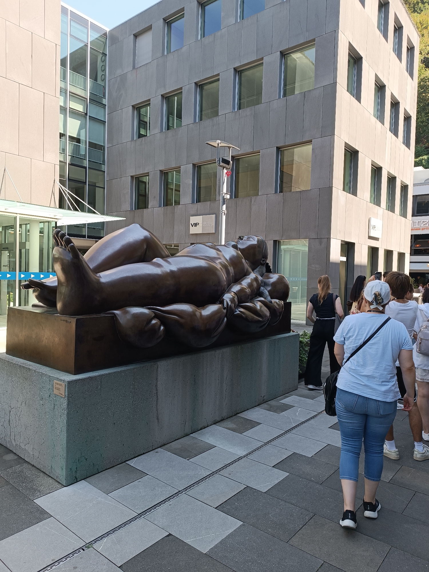 In Vaduz, Liechtenstein, a large bronze sculpture of a reclining figure is displayed on a concrete pedestal. The sculpture depicts a stylized, rounded human form with exaggerated features, seemingly resting. The scene takes place outdoors on a paved walkway adjacent to modern buildings. Several people are visible in the background, walking away from the sculpture, some appearing to be tourists or shoppers. The architecture features large glass windows and stone facades, suggesting a commercial or administrative area. The overall impression is of an urban public space with a significant art installation.
