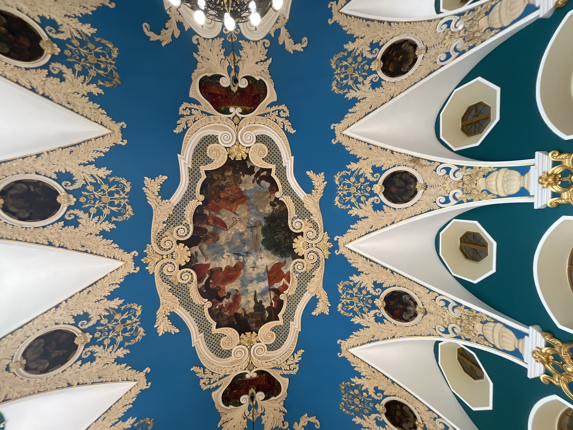 This image shows a section of an ornate ceiling, likely indoors. In the center, a large, framed painting with an elaborate gold border depicts a scene with numerous figures, flags, and what appears to be aerial vehicles, possibly referencing historical events. The painting is set against a blue background. Surrounding this central painting are decorative elements, including white triangular shapes with golden details, and what seem to be other painted ovals. The architecture is reminiscent of classical or Baroque style with intricate details and embellishments. There's also a visible chandelier with multiple lights at the top. The scene gives the impression of being in a grand, historic building, possibly in Moscow, Russia.