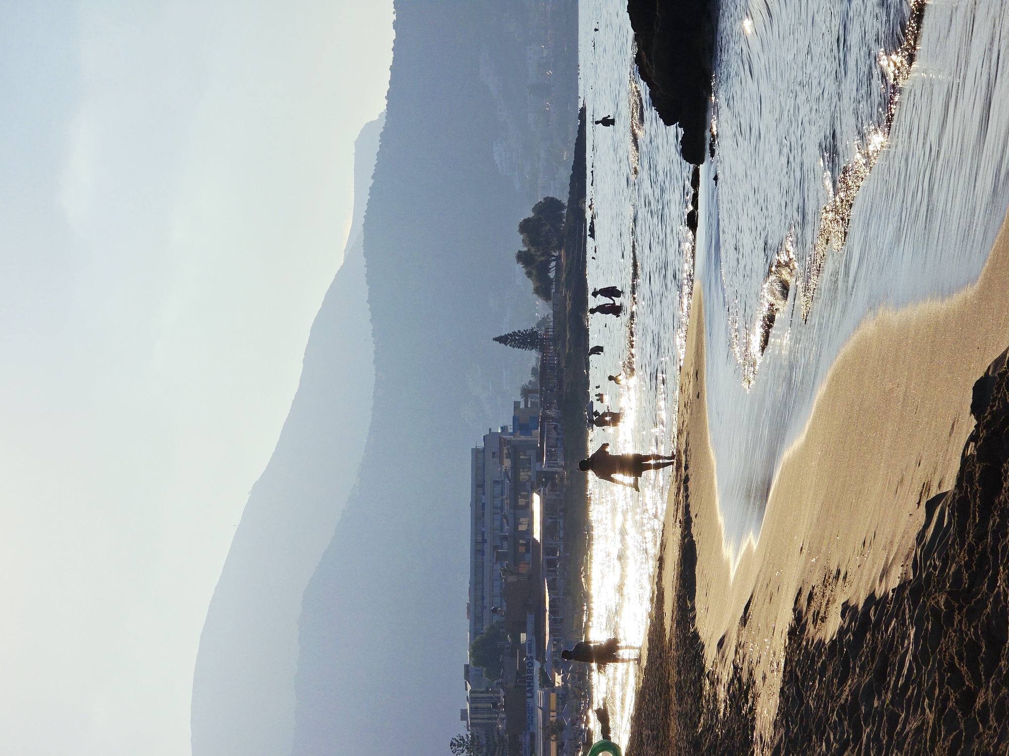 This is a photograph of a beach in Malia Municipal Unit, Greece. The sun is shining, and the water is calm with gentle waves. There are several people in the water, swimming and wading. Some people are walking on the beach, and there are buildings in the background, suggesting a coastal town. The overall scene is one of a relaxed and pleasant summer day at the beach.