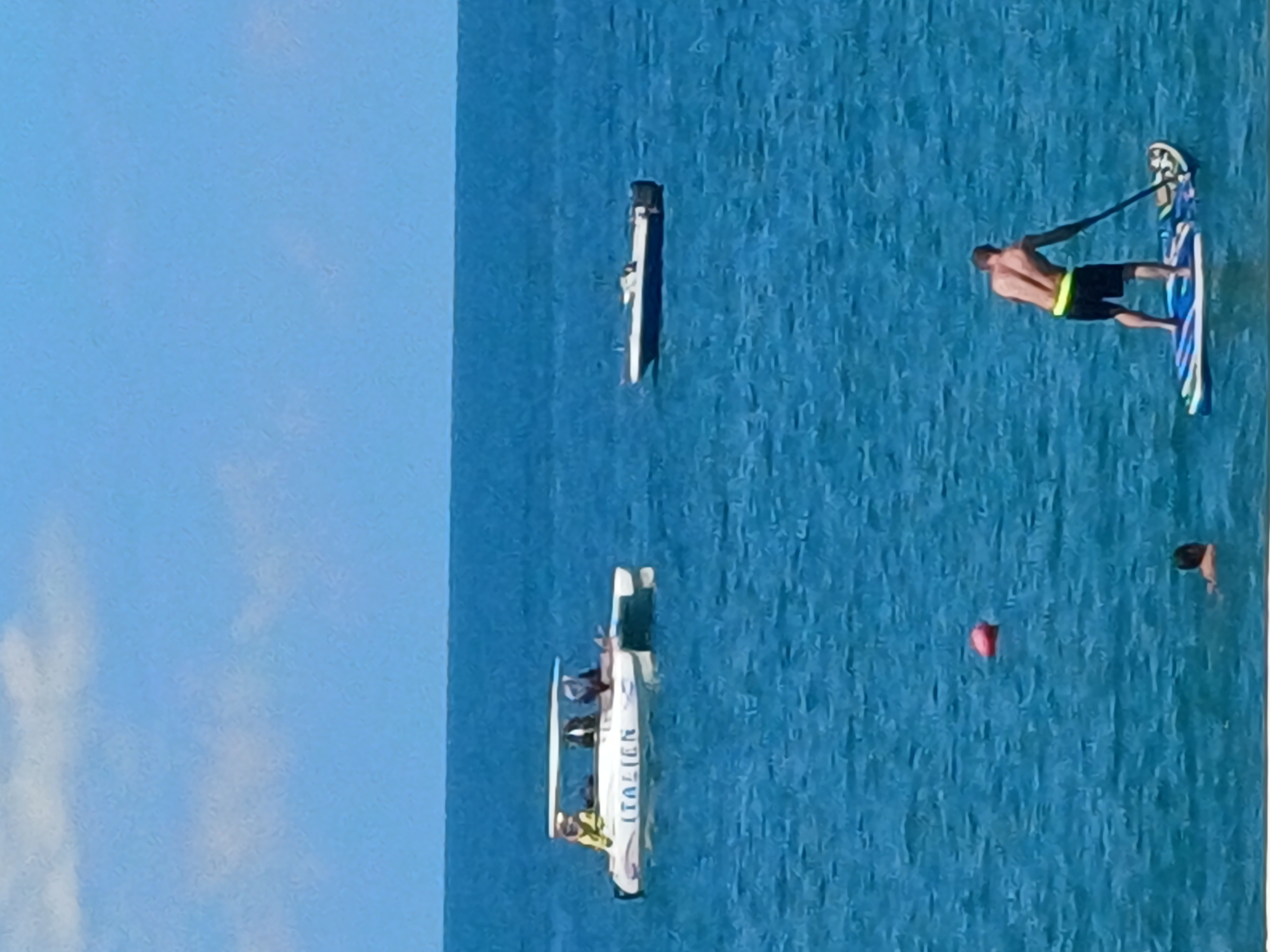The image depicts a wide shot of the Black Sea, likely near Sochi, Russia, under a bright, clear sky with scattered clouds. In the middle distance, a white catamaran with the word "ITALIAN" visible on its side is anchored, with at least three people on board. To the right of the catamaran, a smaller white boat with a dark hull is also at anchor. Closer to the foreground, a man is paddleboarding, facing away from the camera. He is wearing black shorts and a yellow band around his waist. To his left and further back, a red buoy floats on the surface of the water, and a person's head and shoulders are visible in the water. The water is a deep blue with small waves.