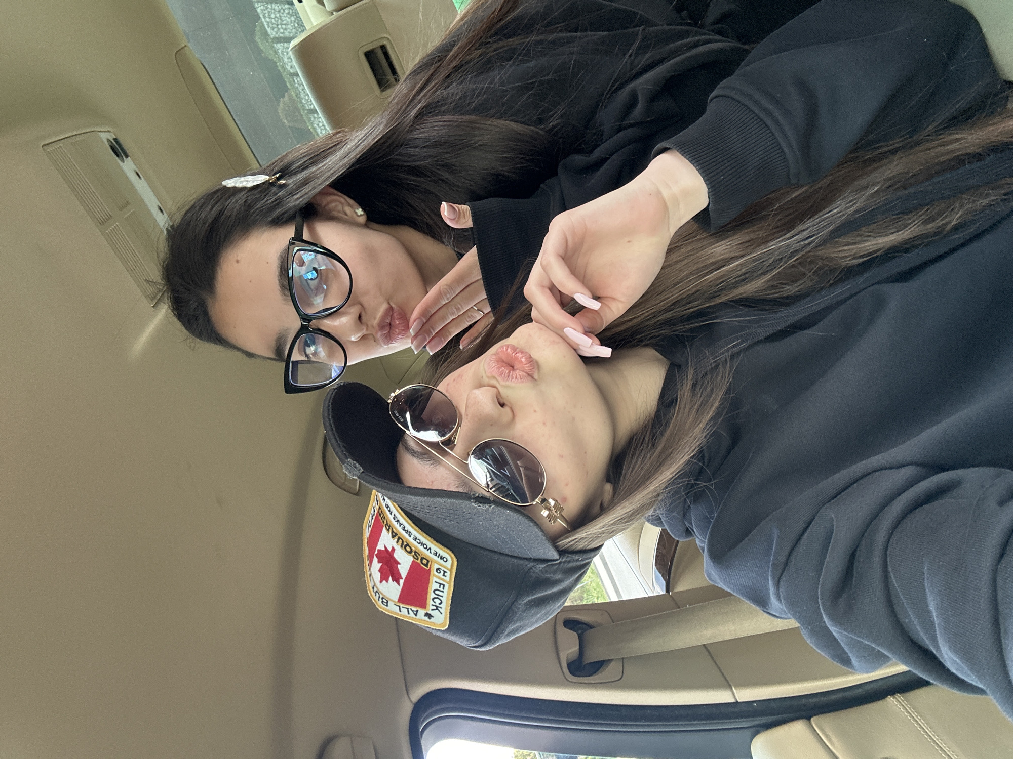 Two women are posing for a picture in what appears to be the interior of a vehicle. The first woman, positioned at the top of the frame, is wearing black framed glasses and has a hair clip in her hair. The second woman, located towards the bottom of the frame, is wearing sunglasses and a black baseball cap with a patch featuring a maple leaf and text. Both women are pouting their lips in a kiss gesture toward the camera. The interior of the vehicle is beige. The location is unknown.
