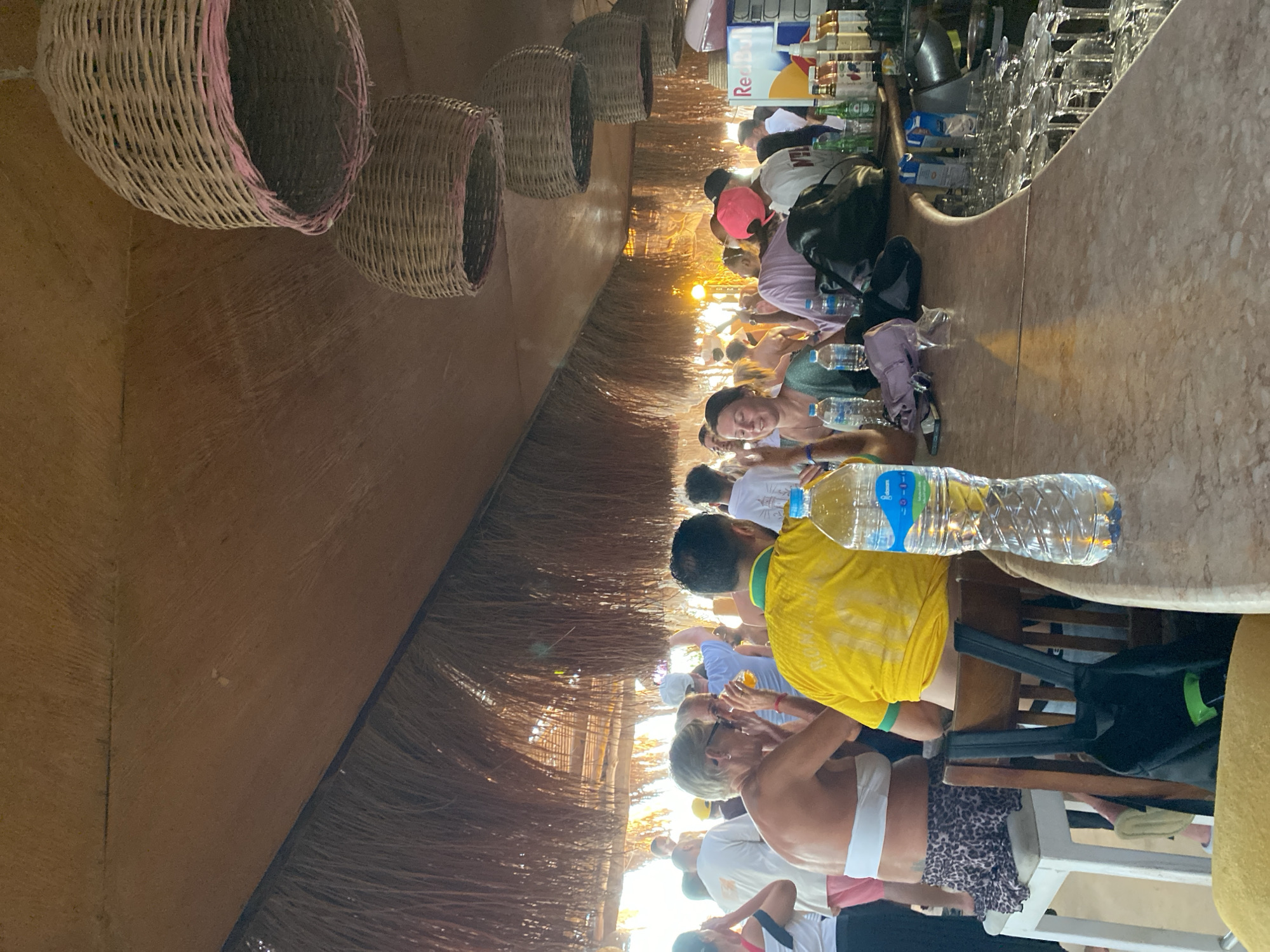 This image shows a group of people sitting at a bar or counter. The setting appears to be outdoors, possibly at a beach bar or a resort, with woven baskets hanging overhead and a thatched roof structure. The people are dressed casually, suggesting a relaxed atmosphere, and some are wearing brightly colored clothing, including a yellow t-shirt and a pink hat.
The activity taking place seems to be socializing and enjoying drinks at the bar. Several water bottles are visible on the counter, along with an assortment of glasses stacked behind the bar. A Red Bull cooler is also present, indicating a selection of beverages. The lighting suggests it is daytime, with sunlight filtering through the roof, creating a warm ambiance.
Notable details include the tropical or beach-like environment due to the thatched roof and baskets. The people's attire and relaxed postures contribute to a sense of leisure and enjoyment. The image captures a moment of social gathering, possibly during a vacation or outing in a warm climate. The overall mood seems cheerful and convivial. There is text visible on a Red Bull cooler, and on some of the water bottles.