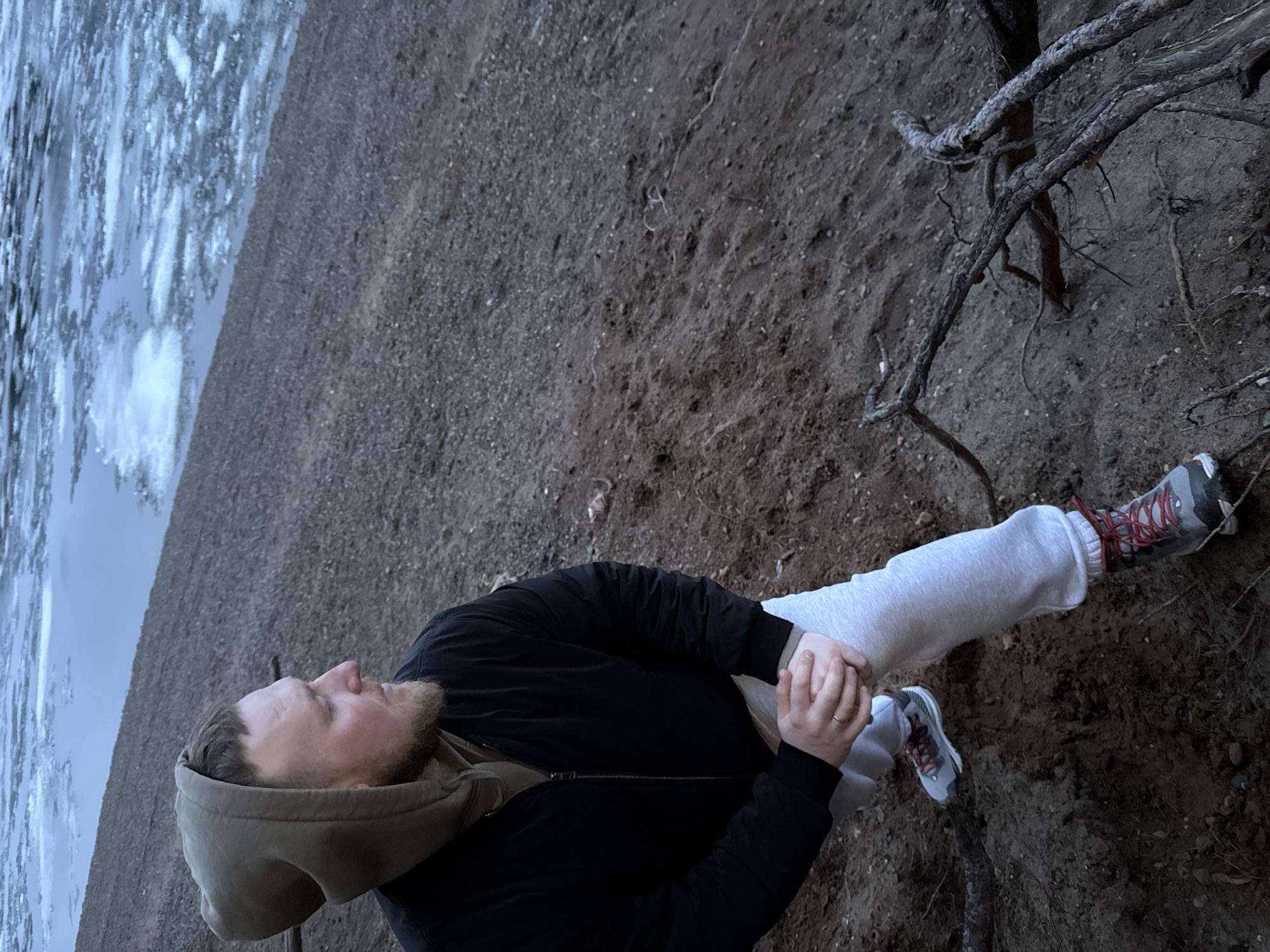 An outdoor scene depicts a bearded man standing on a dirt bank beside a body of water partially covered with ice. The man wears a dark jacket over an olive-green hooded sweatshirt, light grey sweatpants, and light-colored sneakers with red laces. He is looking upwards and towards the icy water, with his hands clasped together in front of him, one hand displaying a ring. The way the hands are clasped suggests he might be holding another person's hand, though only a portion of the second hand is visible.
The ground is composed of dark, coarse dirt and small stones, with several gnarled, bare tree roots and branches extending across the bank to the man's right. The water, visible to the left, is covered with broken ice floes and patches of open water, reflecting the overcast sky. The lighting is subdued, suggesting either an overcast day or early evening. The cool color palette of blues, greys, and browns emphasizes the cold environment, consistent with the presence of ice. The setting appears to be a natural, undeveloped area, possibly a riverbank or lakeside. No text is visible in the image. The man's posture and gaze convey a contemplative or observant mood.