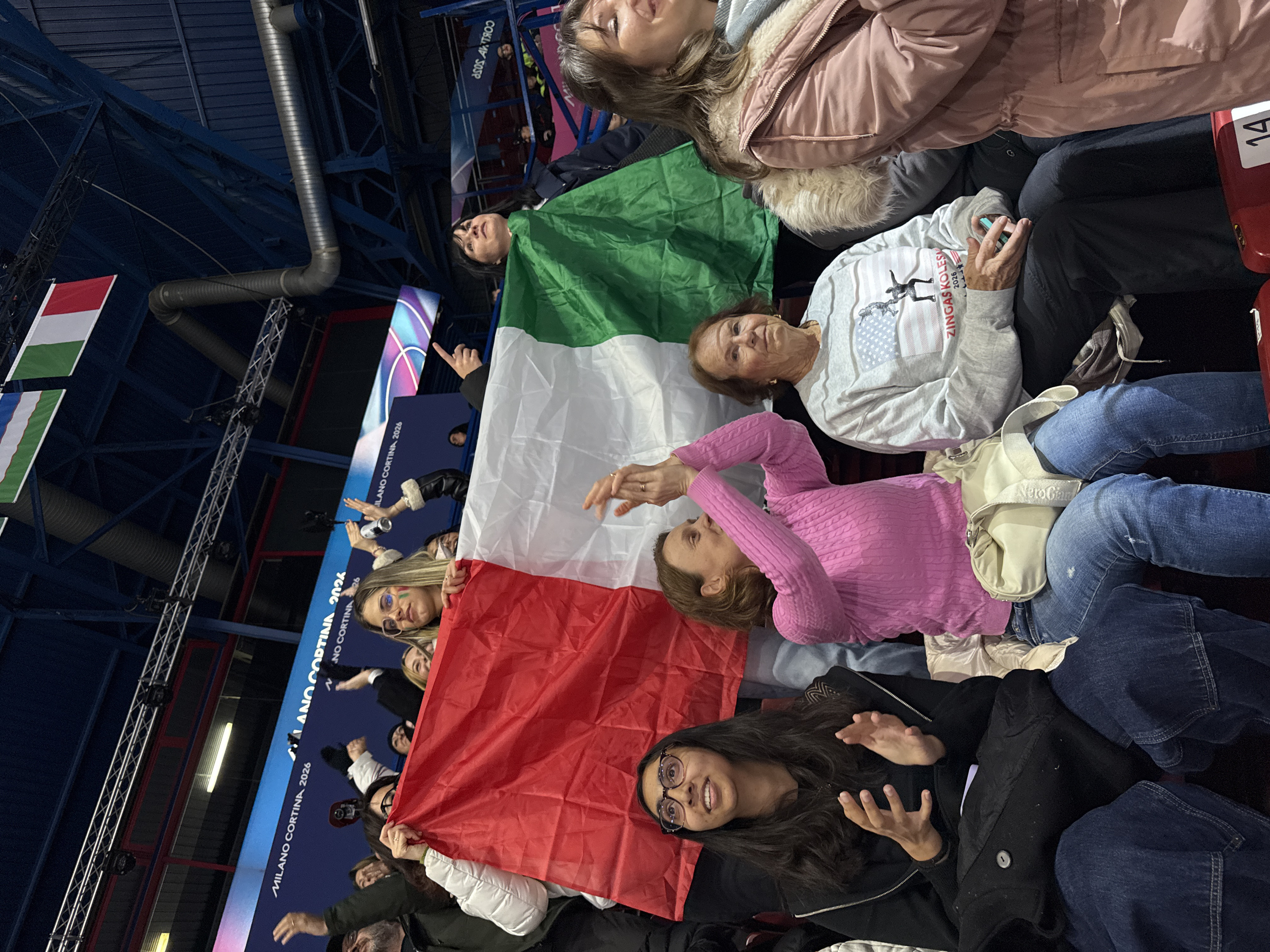 A group of people are seated in stadium seating, holding an Italian flag. The flag's colors are arranged in horizontal bands of green, white, and red. Some individuals are gesturing with their hands, while others are looking upwards. The environment features exposed ceiling structures, along with promotional displays for "Milano Cortina 2026." Additionally, a Hungarian flag is visible.