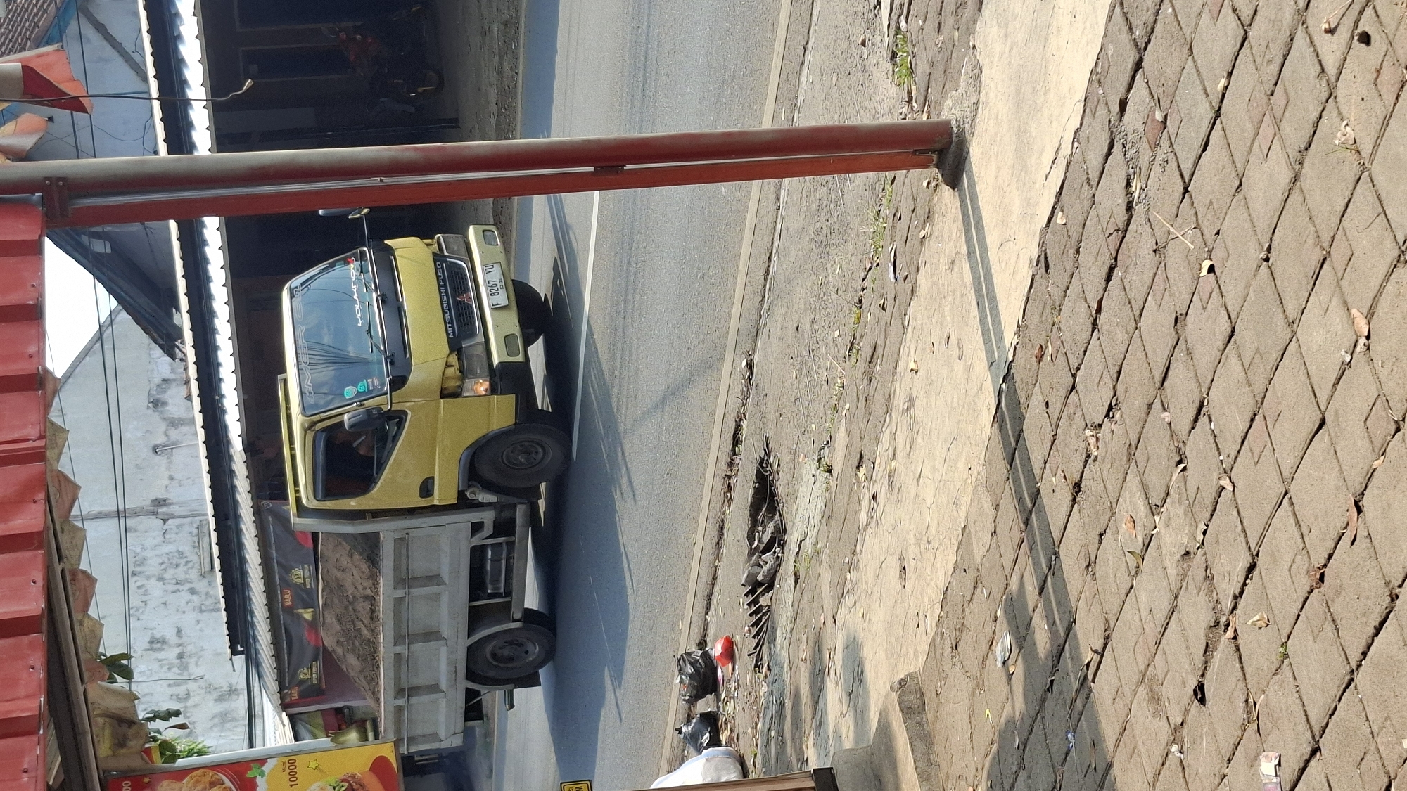 A yellow Mitsubishi Fuso truck is driving on a road in Cilaku, Indonesia. The truck bed is full of dirt or sand. A person is visible in the driver's seat. The truck has a license plate that reads "F 8267 YU". In the foreground, a sidewalk is visible with scattered leaves and a metal grate. Trash bags are also present on the side of the road. In the background, buildings with white walls and dark roofs are visible, as well as trees and a red pole. The scene appears to be a street in a commercial or residential area.