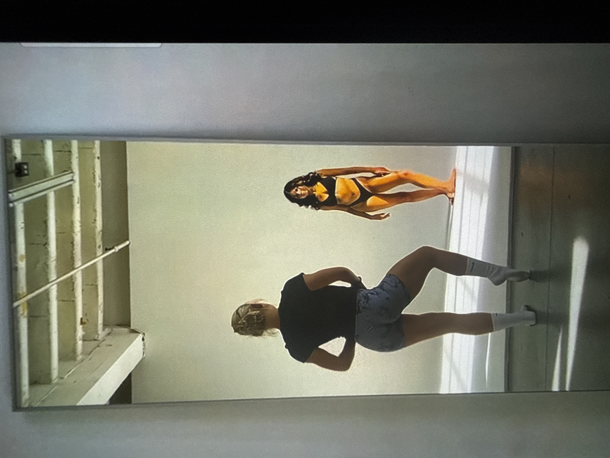 The scene depicts a large mirror reflecting two women within what appears to be a studio located in Varna, Bulgaria.
In the foreground reflection, a woman with blonde hair styled in a bun is viewed from behind. She wears a black short-sleeved top, patterned grey athletic shorts, and white crew socks, with her left foot flat on the floor and her right foot on pointe. Her body is angled, and she is observing her own reflection and the other woman in the mirror.
In the background reflection, a woman with long dark hair stands facing forward, wearing a black two-piece bikini comprising a sports bra-style top and high-cut bottoms. She is barefoot and illuminated by a strong, warm-toned light, casting distinct shadows behind her on the white surface she stands upon.
The reflected environment reveals a high ceiling with exposed white beams and concrete blocks, along with visible conduit or piping. The studio walls are plain and light-colored. The floor, where the woman in shorts stands, is a light grey or concrete, marked by sharp, elongated shadows from an unseen light source. The overall impression is a studio setting, possibly for a photoshoot or dance practice, with both individuals engaged in posing or physical activity.