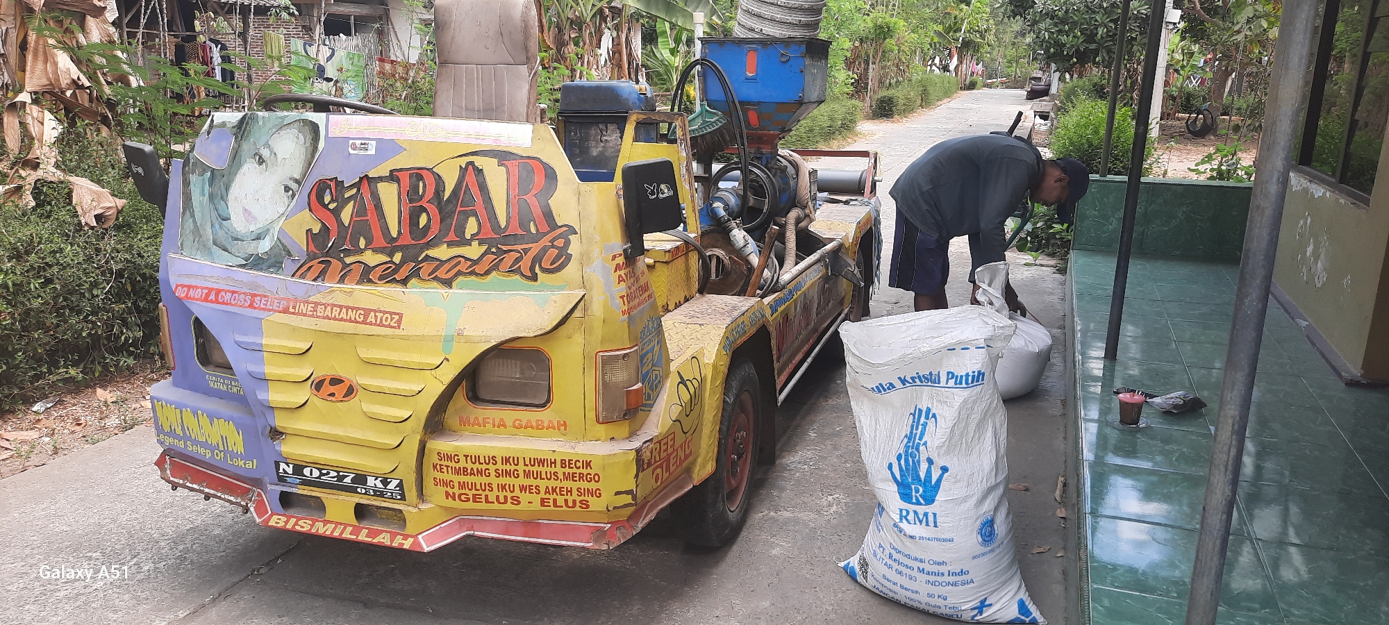 In Ploso, Indonesia, a man wearing a dark long-sleeved shirt and dark shorts is interacting with large white sacks on a concrete path. He is bent over, appearing to be handling or organizing the sacks. Two large, white sacks are prominently displayed, one with blue lettering that includes "Gula Kristal Putih" and the RMI logo. To the left, a yellow vehicle is parked, decorated with various decals and text. The vehicle's rear features license plate "N 027 KZ 03-25" and the word "BISMILLAH" in red. The vehicle has a faded depiction of a woman wearing a hijab on its left side. The environment includes dense greenery and a paved road in the background. The overall scene suggests an outdoor setting, possibly a roadside or a yard, related to the handling of goods.