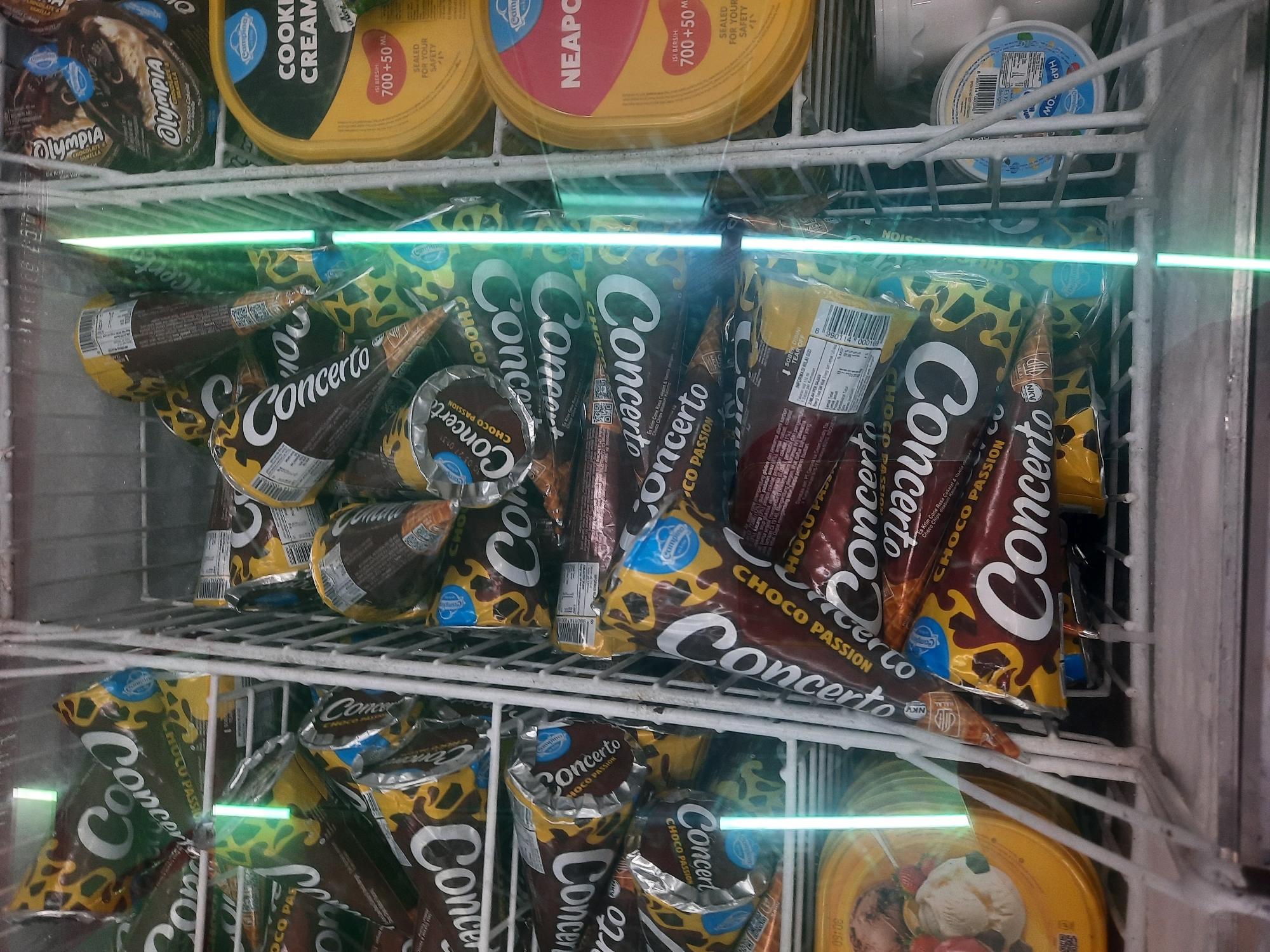 The image is a close-up, eye-level, interior shot of a commercial freezer display case filled with various ice cream products, primarily ice cream cones. The products are organized in wire racks within the freezer.
The dominant items are "Concerto" brand ice cream cones, with packaging featuring brown, yellow, and leopard print designs. The cones are stacked and arranged in multiple layers. Some individual cones are visible, showcasing their wrappers, which also bear the "Concerto" branding and "Choco Passion" flavor.
To the right of the "Concerto" cones, there are round tubs of ice cream. One is yellow with a black and red label that reads "Campina COOKIE CREAM". Another tub, to its right, is also yellow with a prominent red label that says "NEAPOLITAN". Further to the right and in the background, a white tub with a blue and white label is partially visible.
The freezer's interior lighting casts a bright, vertical green glare down the center of the image, obscuring some details of the ice cream products behind it. Frost is visible on the interior surfaces of the freezer and on the wire racks.
The presence of "Campina" and "NEAPOLITAN" brands, and the general context of a freezer display, strongly suggests this is a retail environment like a convenience store or supermarket. Based on the branding of "Campina" ice cream, which is a well-known Indonesian brand, and the prevalence of Indonesian language on some packaging labels (e.g., "Es Krim Rasa Cokelat & Vanila" which translates to Chocolate & Vanilla Ice Cream), this scene is likely located in Indonesia. While the specific city of North Jakarta cannot be definitively confirmed solely from the visual information, it aligns with the geographical context where such products and retail environments are common.
There are no people, actions, or interactions depicted in the image. The scene is static and focuses on the frozen products.