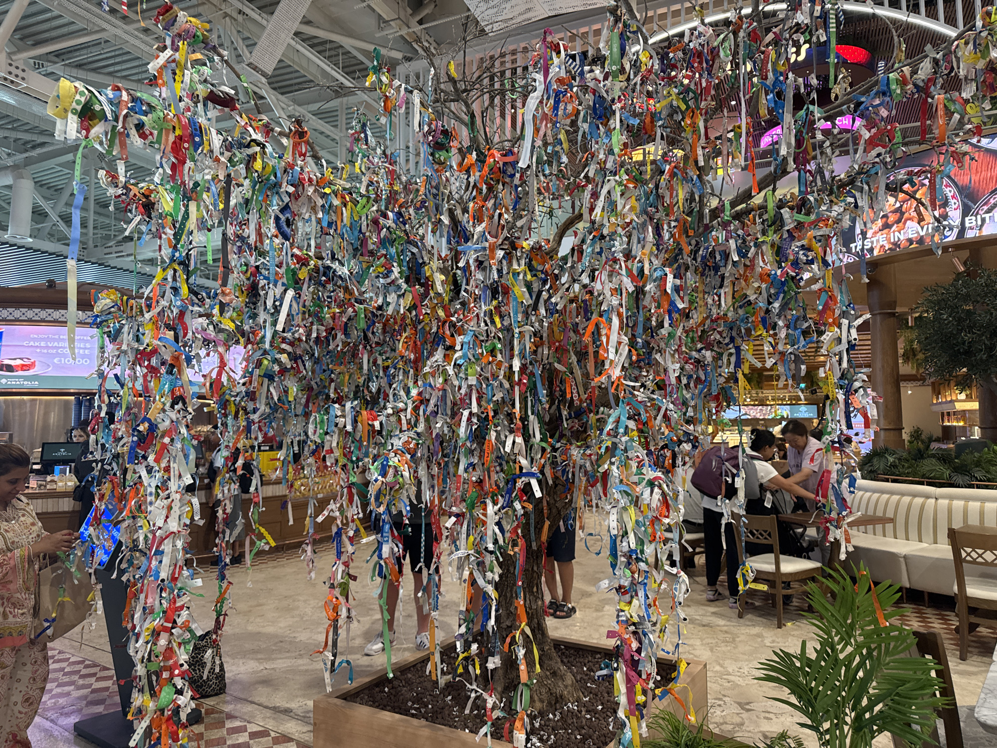 The image depicts a colorful, man-made tree adorned with numerous brightly colored ribbons tied to its branches. The tree is situated indoors, likely in a public space such as a mall or airport terminal, indicated by the presence of retail displays and seating areas in the background. Several individuals are visible in the scene, some interacting with the tree or nearby displays, while others are seated at tables. The overall impression is one of a decorative installation, possibly a wishing tree or an artistic display, placed within a commercial environment. The city of Aksu, Turkey, is not explicitly visible in the image, and its connection to the scene is unclear.