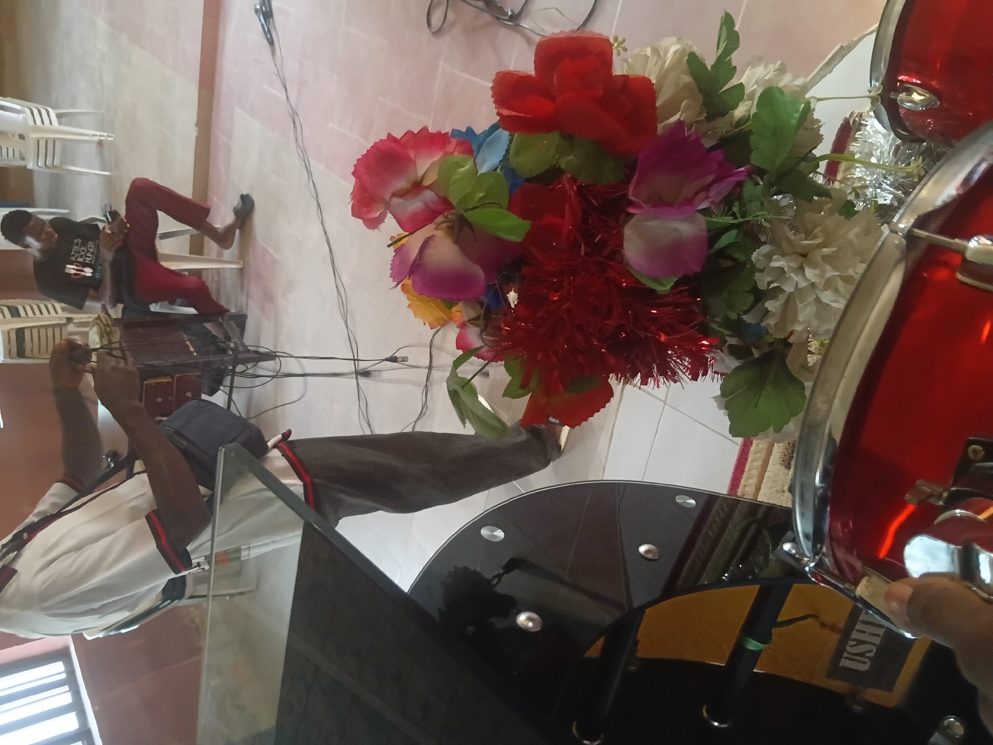 The image depicts an indoor scene with two male individuals and various objects, suggesting a setup for an event, possibly musical.
In the foreground, at the bottom left, the top of two red drums with chrome hardware is visible, partially obscured by a dark, reflective surface, possibly a music stand or table. Below this surface, a yellow panel with the partial inscription "USHE" (likely "USHER") in black text is visible, along with a black microphone stand featuring a green band. A hand is seen near the drums on the bottom left.
Centrally positioned, in the mid-ground, is a vibrant bouquet of artificial flowers, including red, pink, purple, yellow, and white blossoms, intermixed with green leaves and red tinsel-like decorations. Black wires are strewn across the light-tiled floor around the flowers.
To the left of the center, a standing adult male is partially visible from the chest down. He wears a white short-sleeved collared shirt with red and black trim on the collar and sleeves, dark trousers, and a dark shoulder bag. His hands are occupied with black cables, suggesting he is managing equipment wiring. His face is not visible.
To the right, in the background, a younger male is seated on a white plastic chair. He wears a dark short-sleeved t-shirt with a white graphic that partially reads "ARTICS ROCK HUNGER" and dark red trousers. He has a dark slipper on his right foot, while his left foot is bare. He is looking down at a small object held in his hands.
The floor is tiled with light-colored square tiles. The background shows a reddish-brown wall on the left with a window featuring vertical elements, and multiple white plastic chairs stacked on the right. The overall lighting is bright, illuminating the indoor space. No specific visual elements confirm the location as Jalingo, Nigeria.